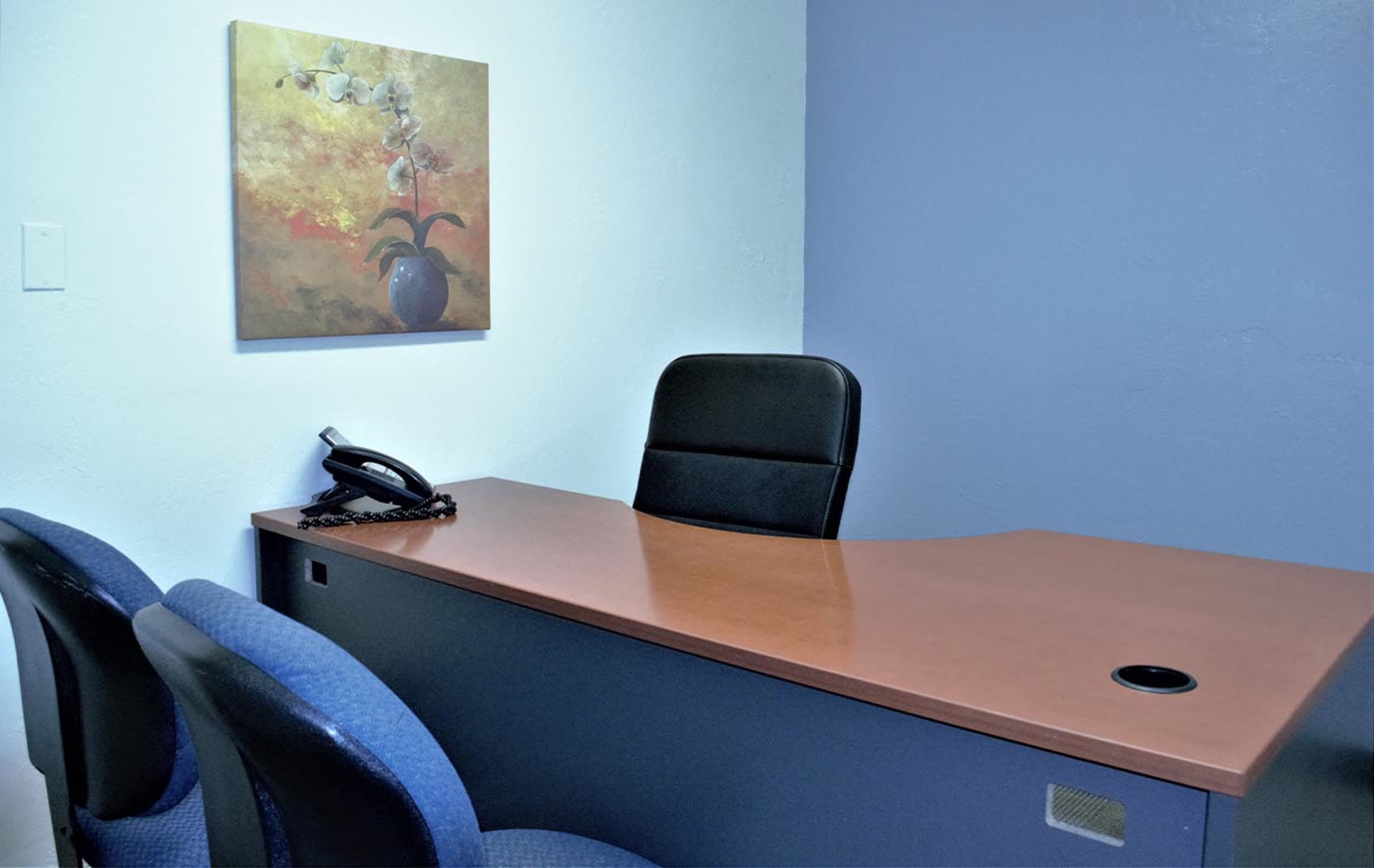 The image shows a simple office space featuring a wooden desk with a phone, three blue chairs, and a floral painting on the wall.
