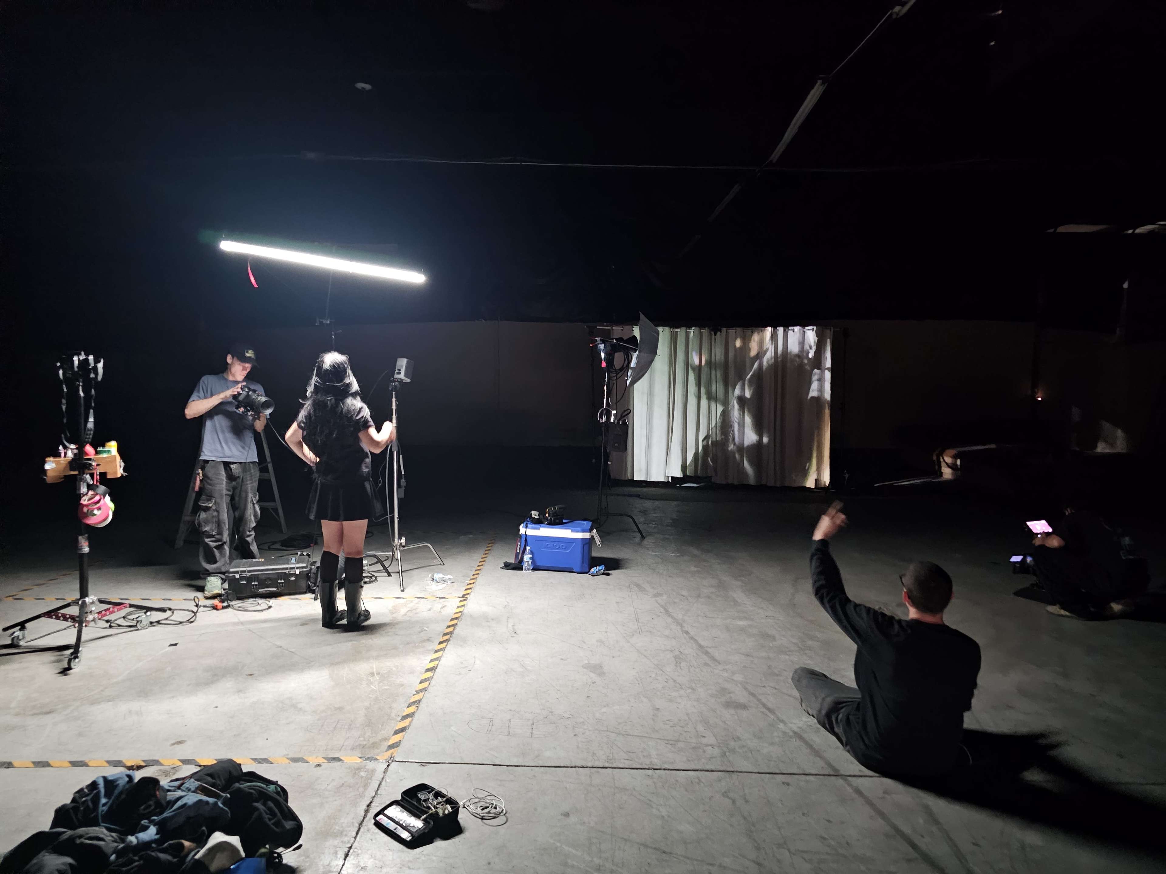 A woman in a black outfit stands in front of a backdrop while two men operate cameras and equipment in a dimly lit warehouse.