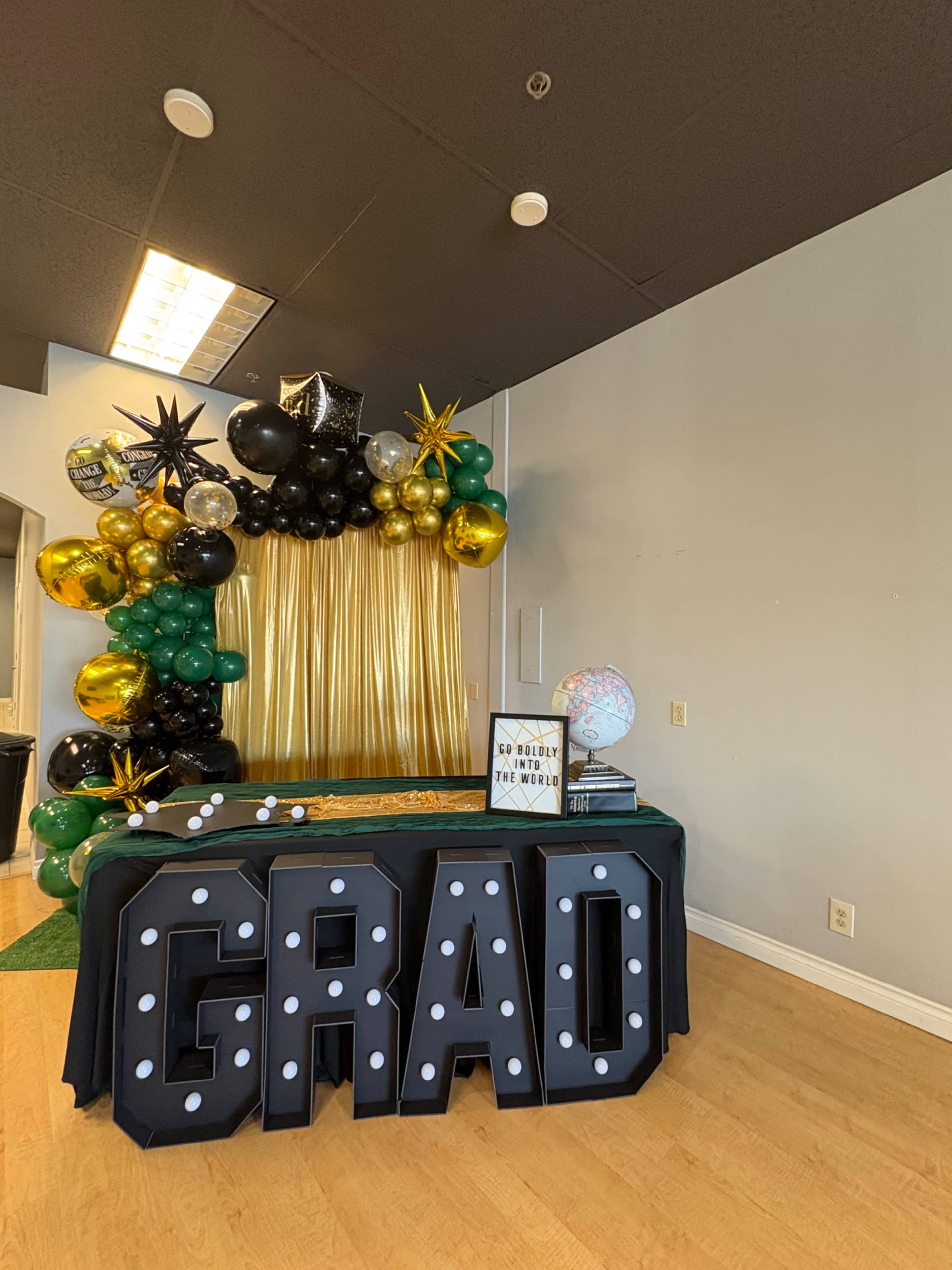The image shows a decorated area for a graduation celebration featuring a gold curtain backdrop, black and green balloons, a globe, and a table with a large "GRAD" sign.