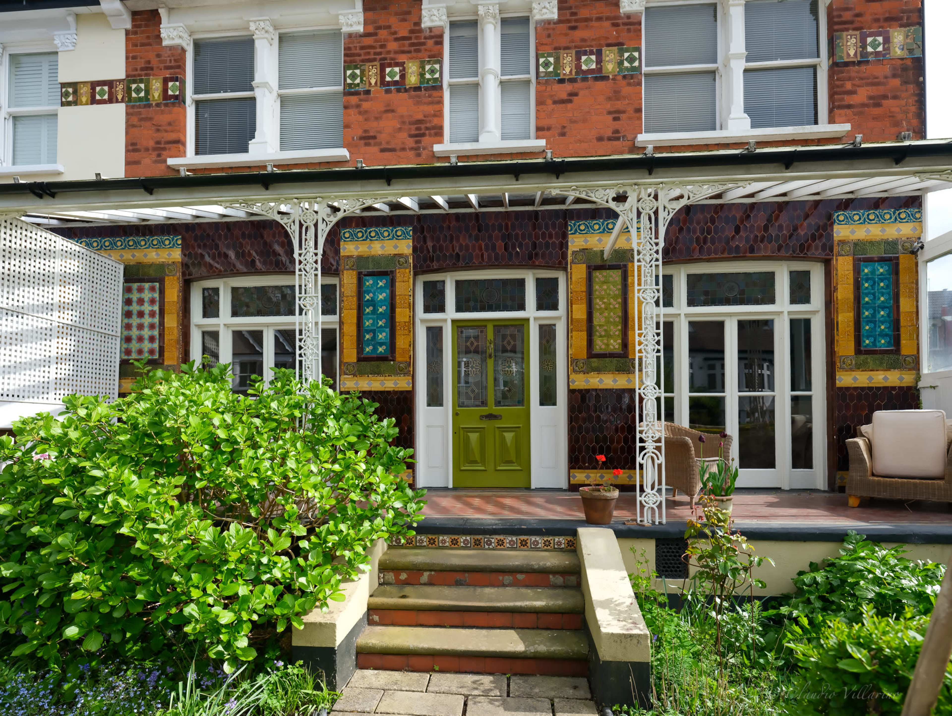 A colorful, intricately decorated facade of a two-story house with a front porch and green door, surrounded by greenery.