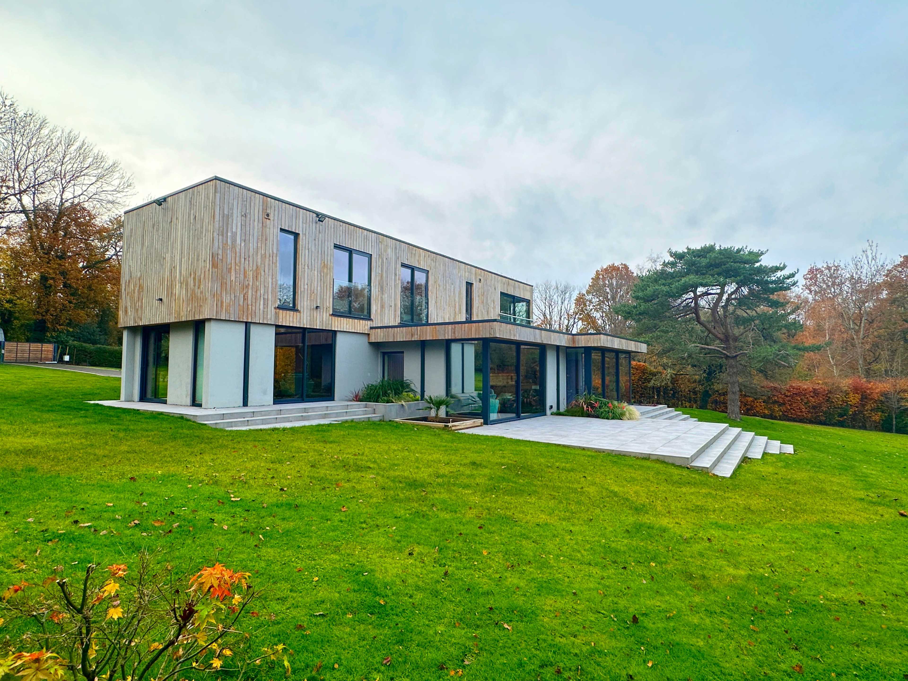 A modern two-story house with a wooden facade and large windows is situated on a green lawn surrounded by trees.