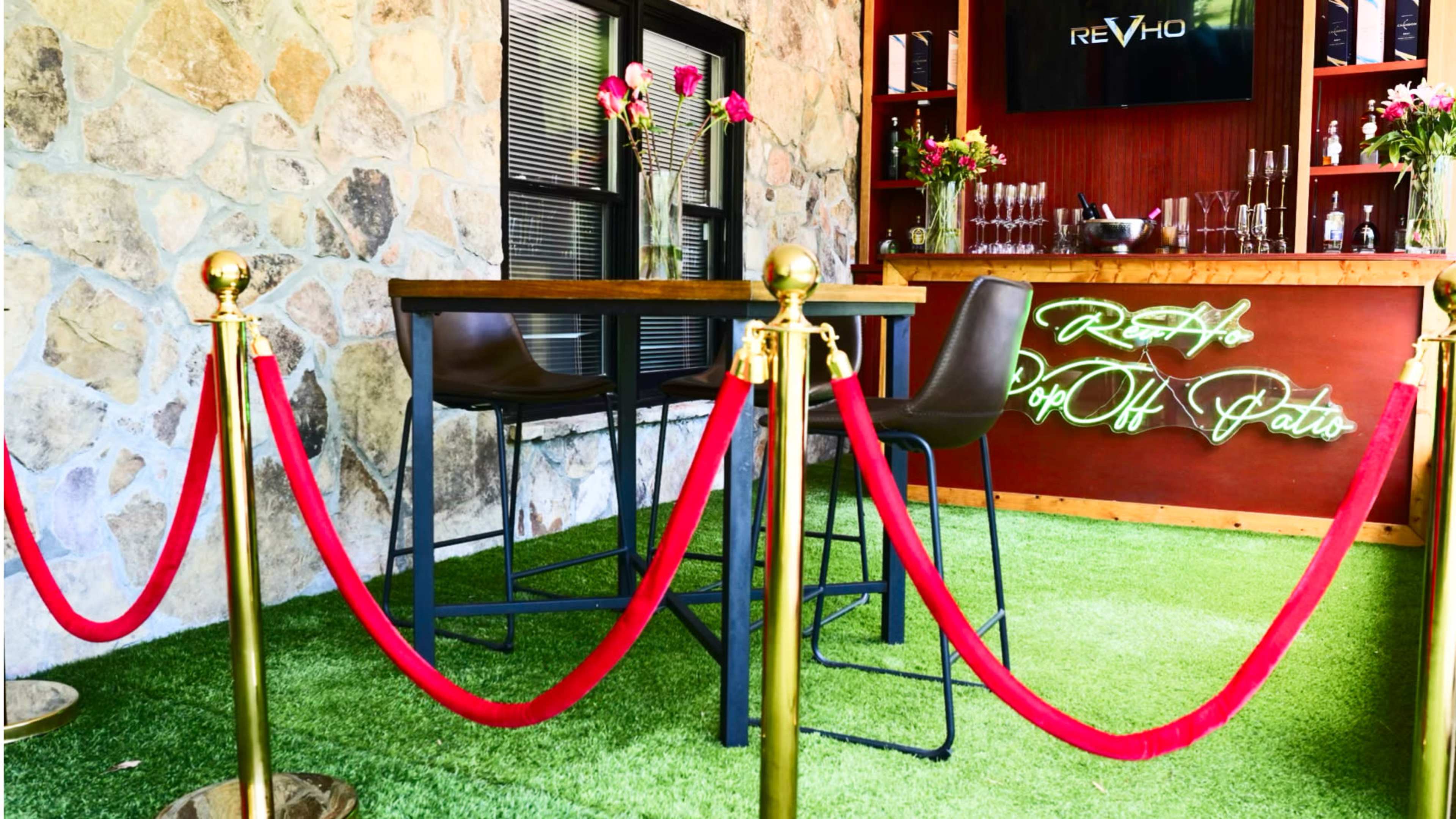 A stylish event space features a wooden table with high stools, surrounded by a stone wall and adorned with flowers, alongside red velvet ropes and a neon sign.