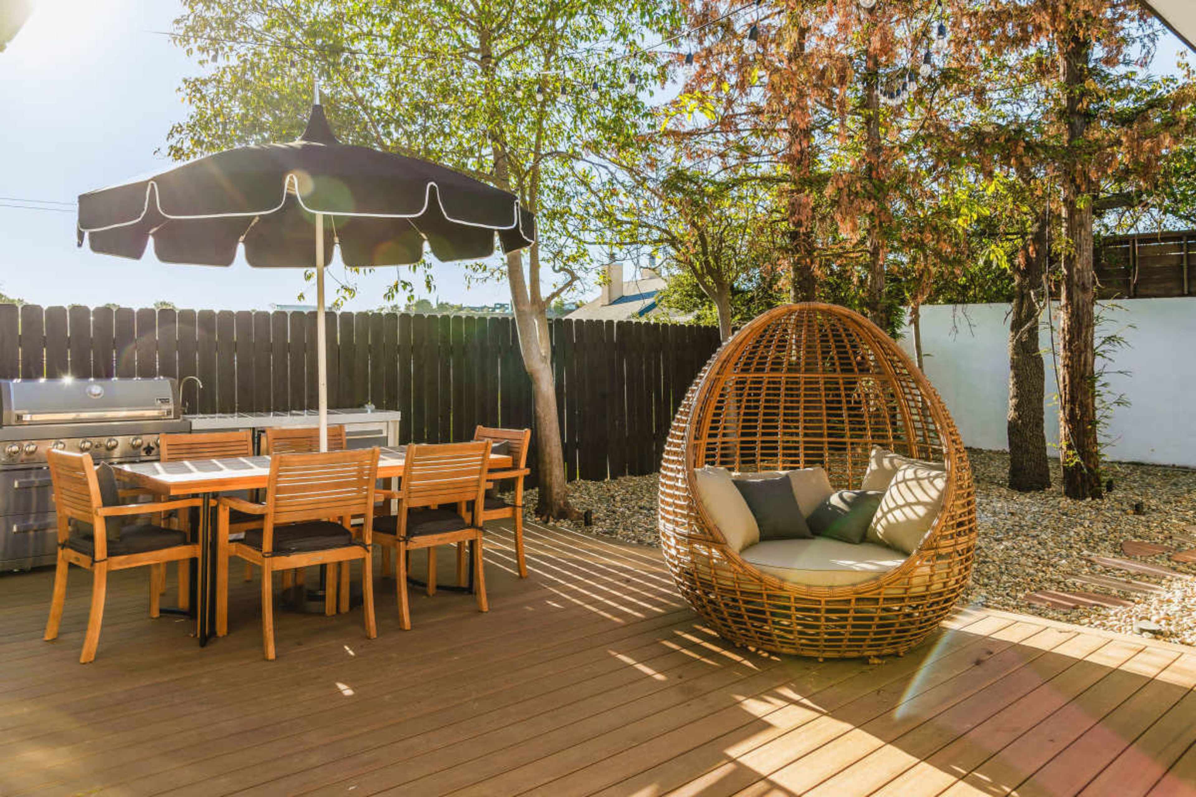 The image shows a backyard patio with a dining set, a large umbrella, and a wicker hanging chair surrounded by greenery and a wooden fence.