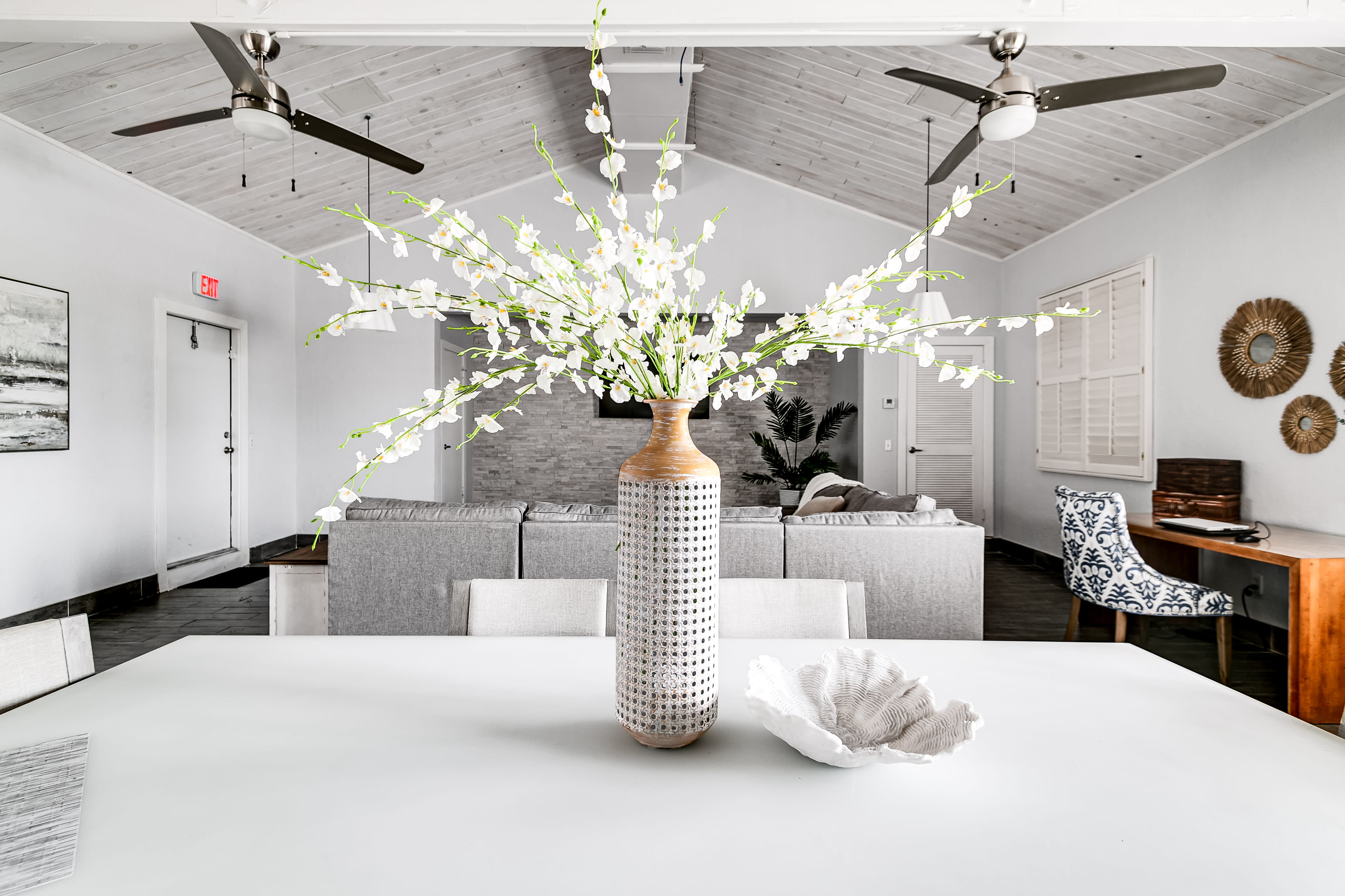 A white dining table with a textured vase filled with white flowers is positioned in a bright, modern living space featuring ceiling fans and neutral-colored furniture.