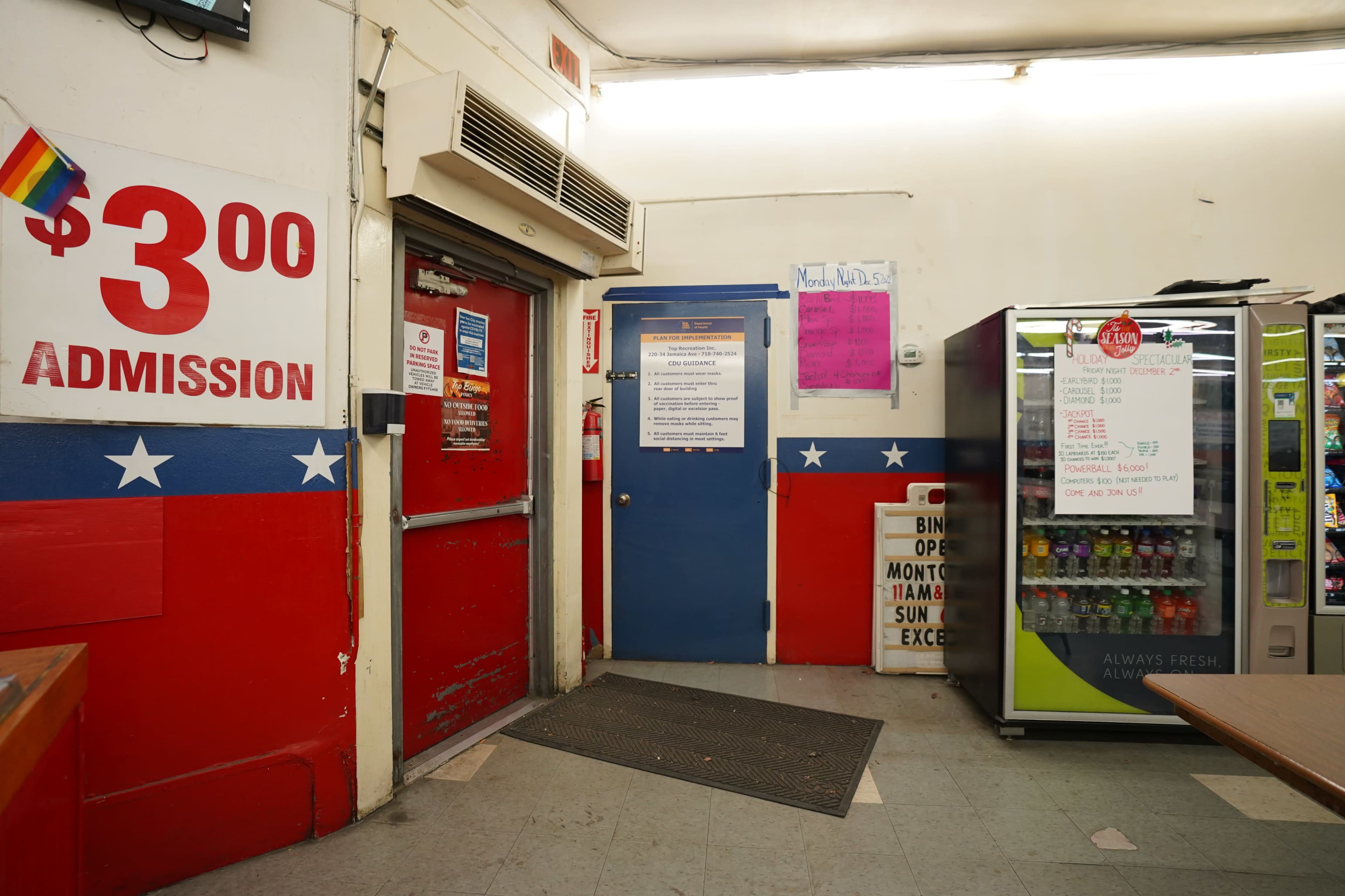 The image shows an interior of a building with red and blue walls, featuring a sign indicating a $3 admission fee and a vending machine in the corner.