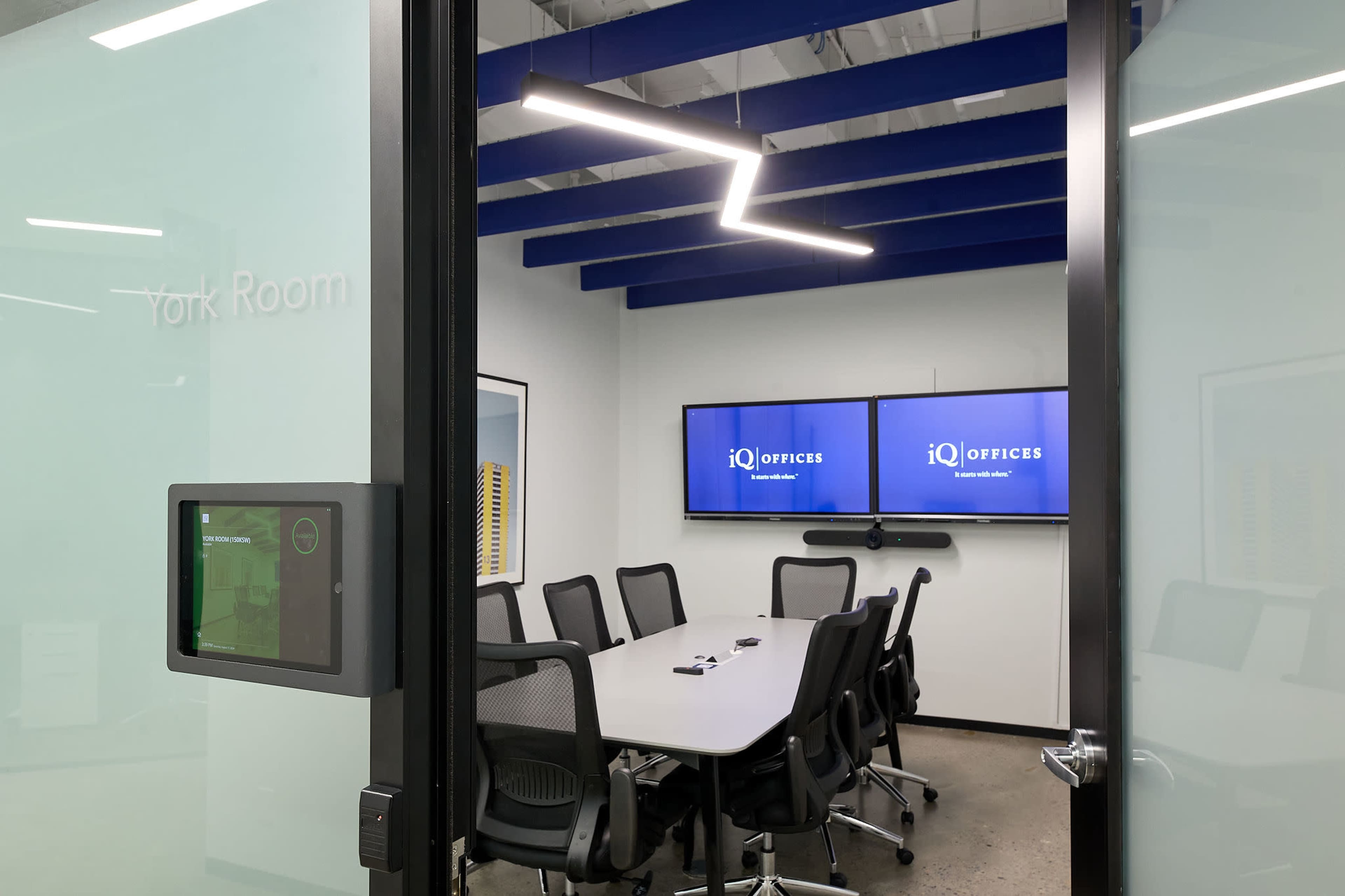 A glass-walled conference room features a large table surrounded by black chairs, with two screens on the wall displaying the iQ Offices logo.