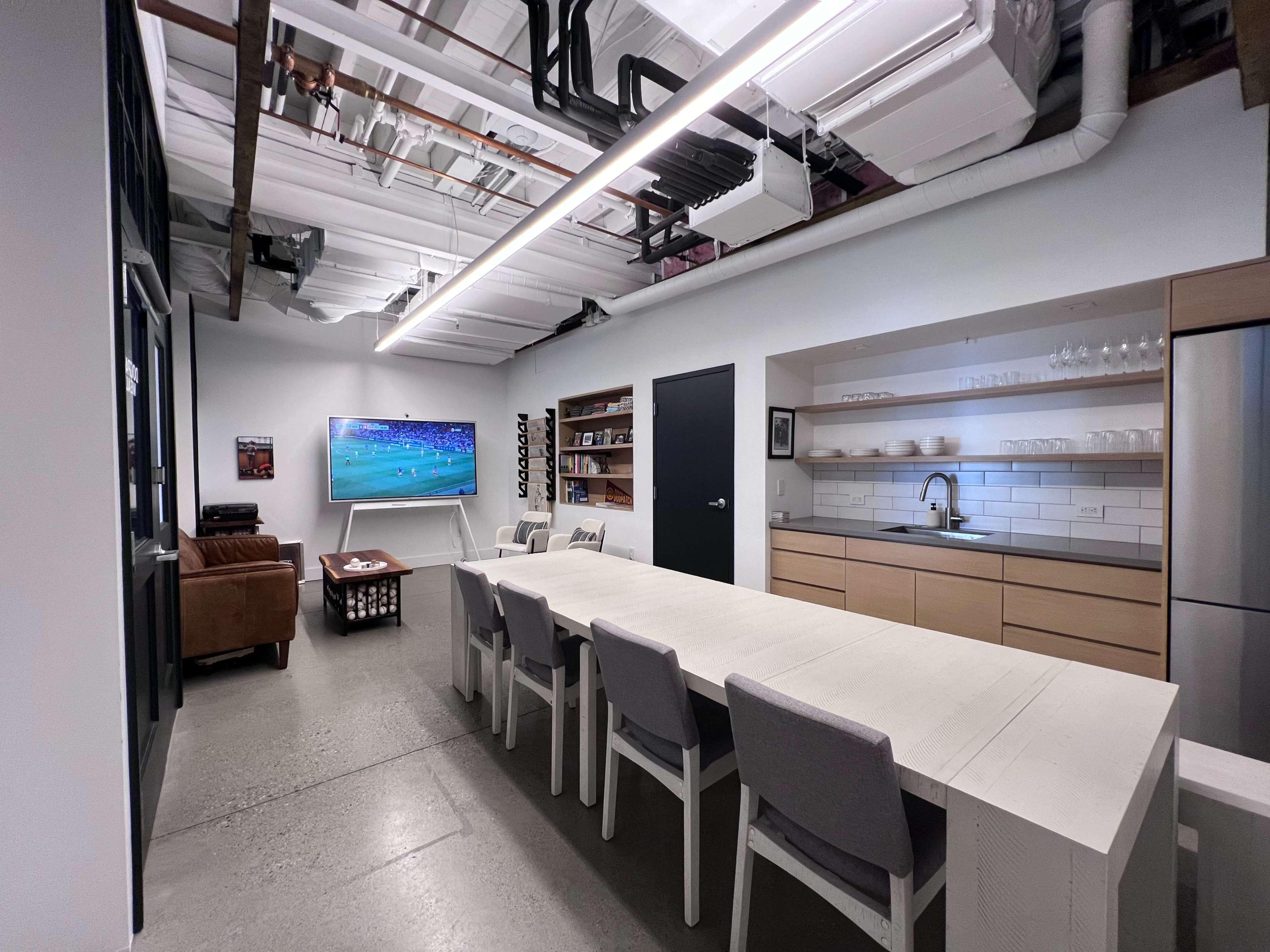 A modern, open-concept room with a long dining table, a television displaying a sports game, a brown sofa, shelving, and a kitchen area with light cabinetry.
