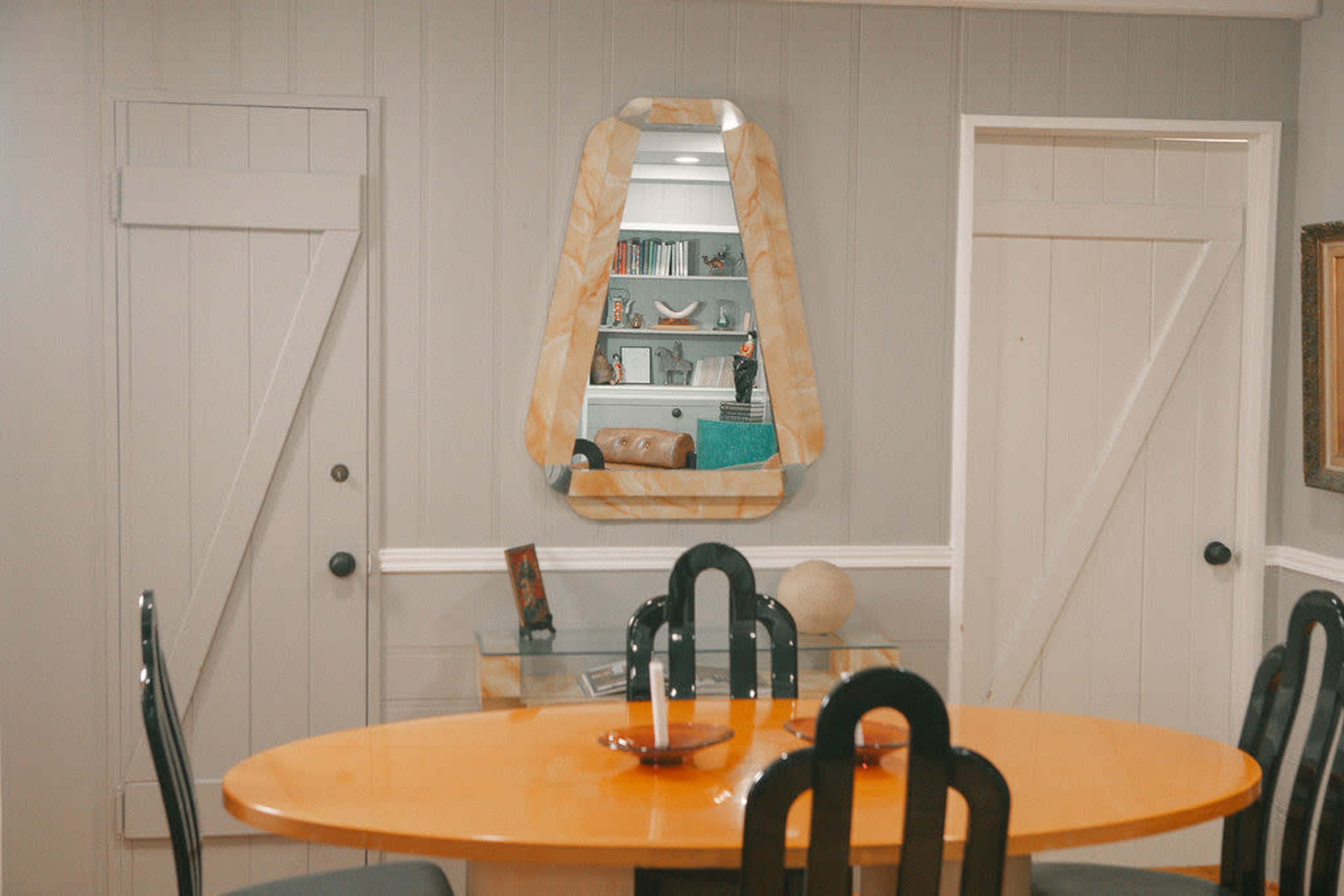 A dining room with an orange circular table, a triangular mirror on the wall, and two doors leading to adjacent rooms.
