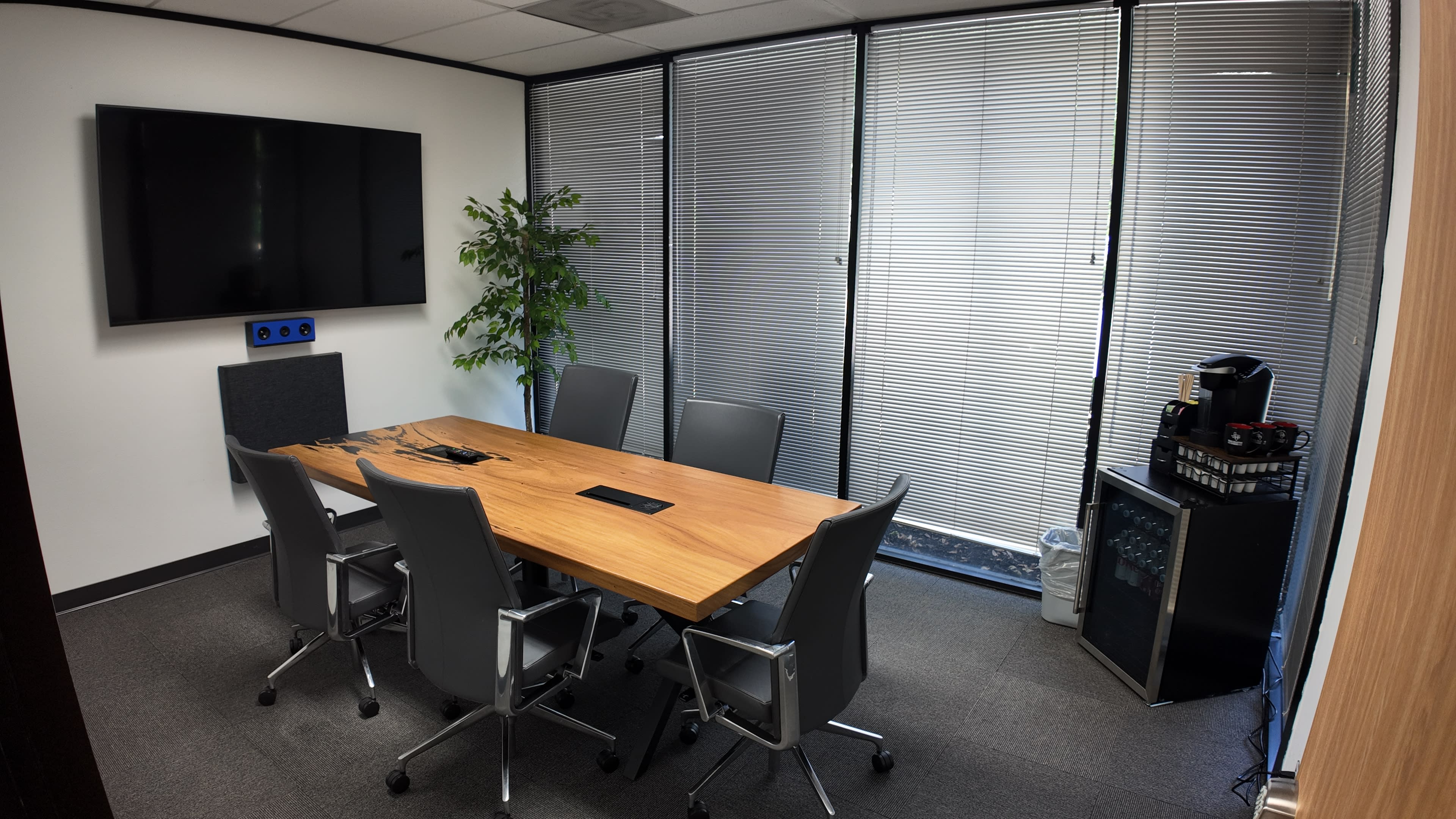 The image shows a conference room with a large wooden table surrounded by six gray chairs, a television mounted on the wall, and a small plant near the window.