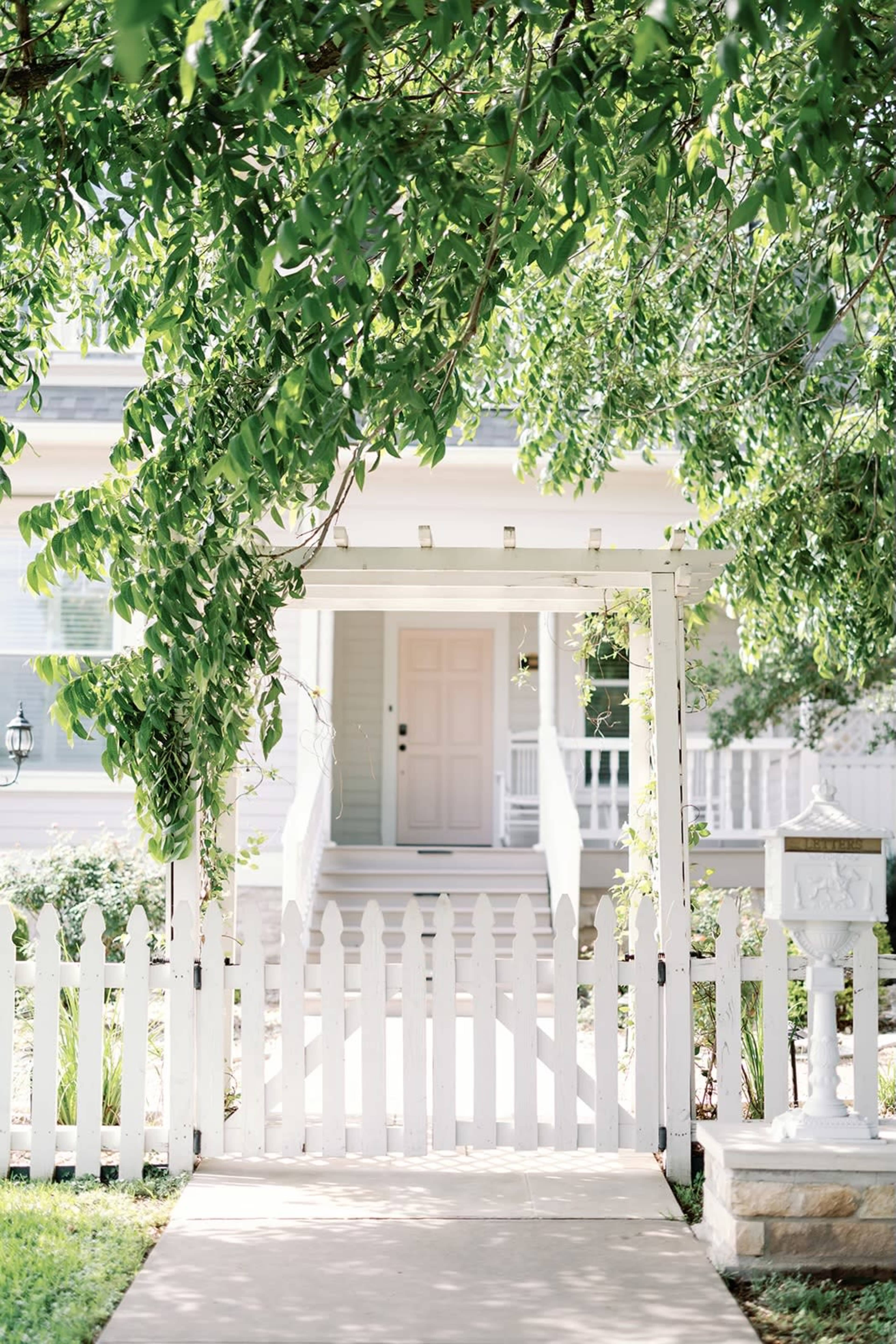 A white picket fence surrounds a pathway leading to a light-colored house with a front porch and a partially shaded entrance.
