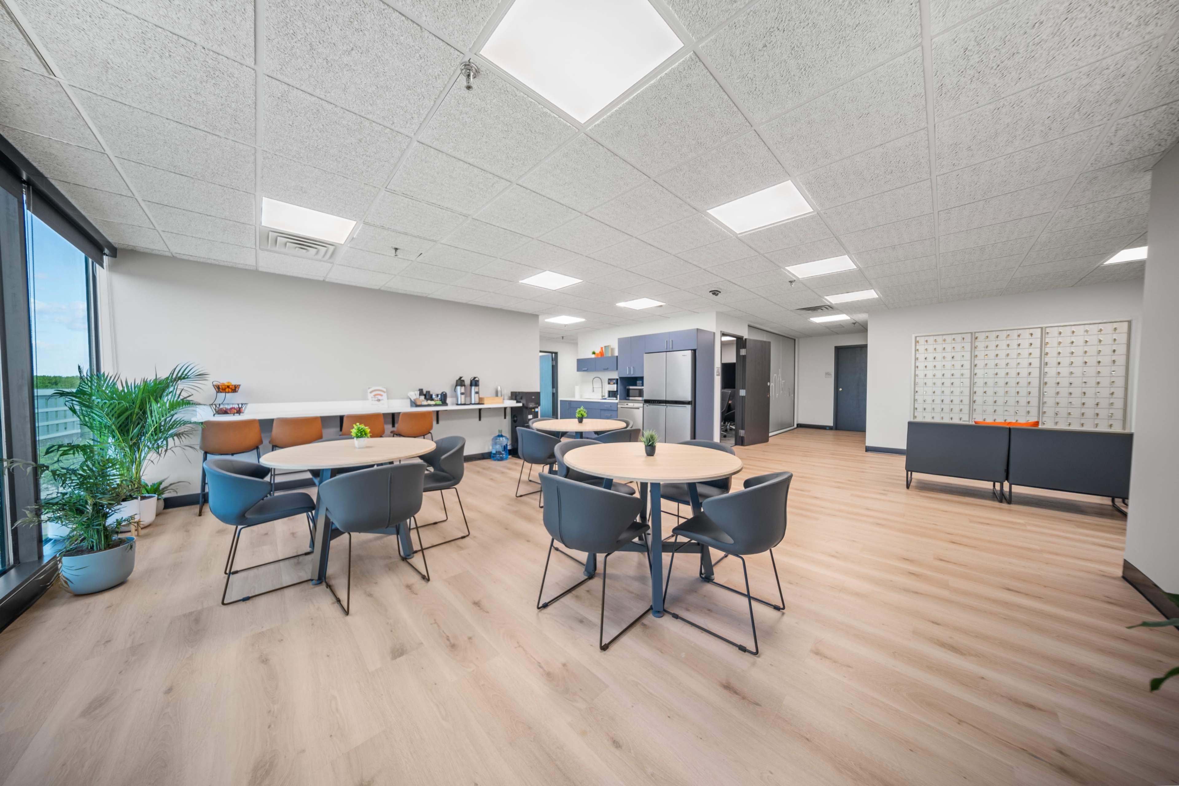 The image shows a modern office break room with two round tables, chairs, and kitchen amenities in a spacious area filled with natural light.