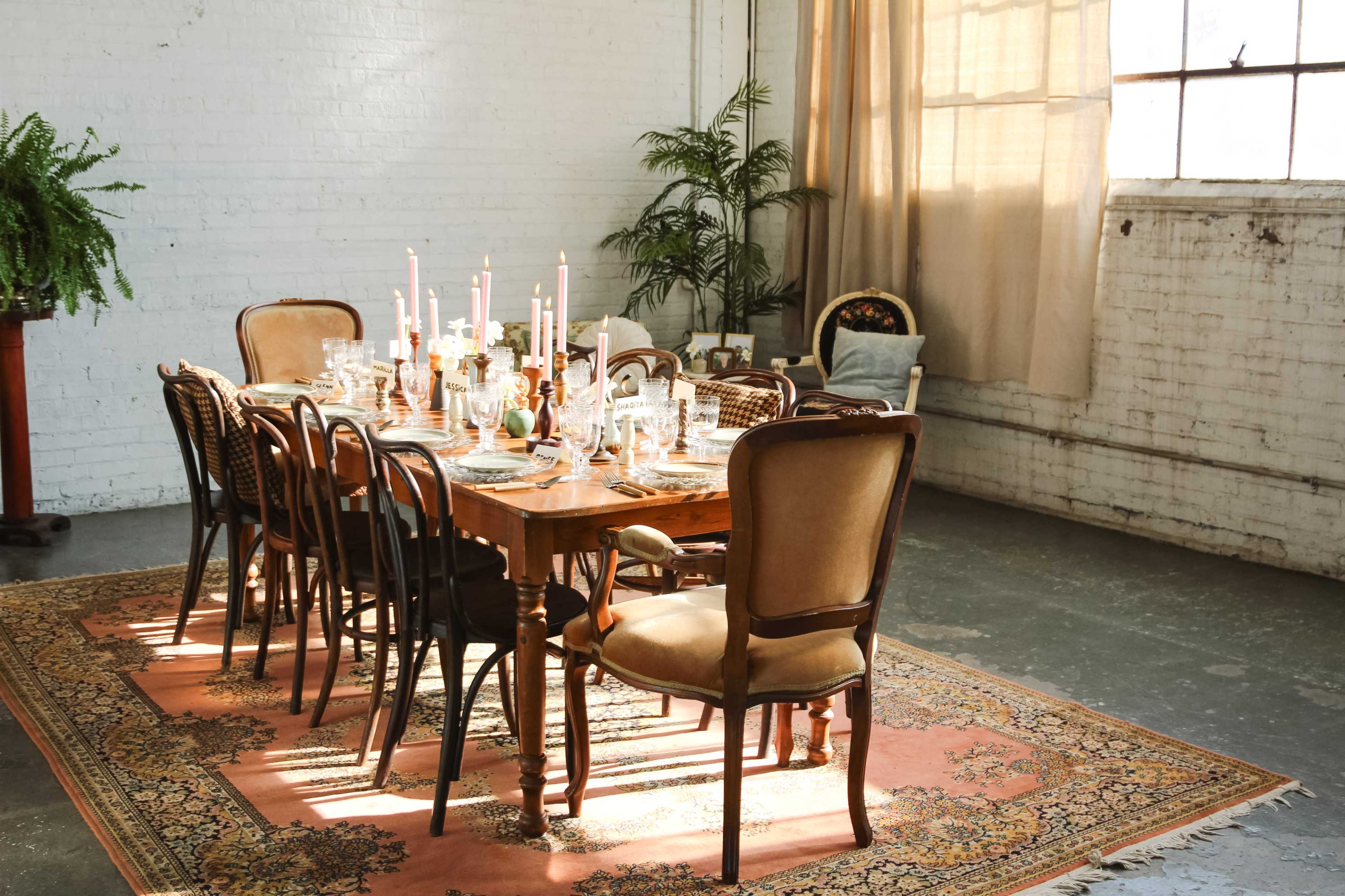 A wooden dining table is set with candles and glassware in a well-lit room featuring a large window and potted plants.
