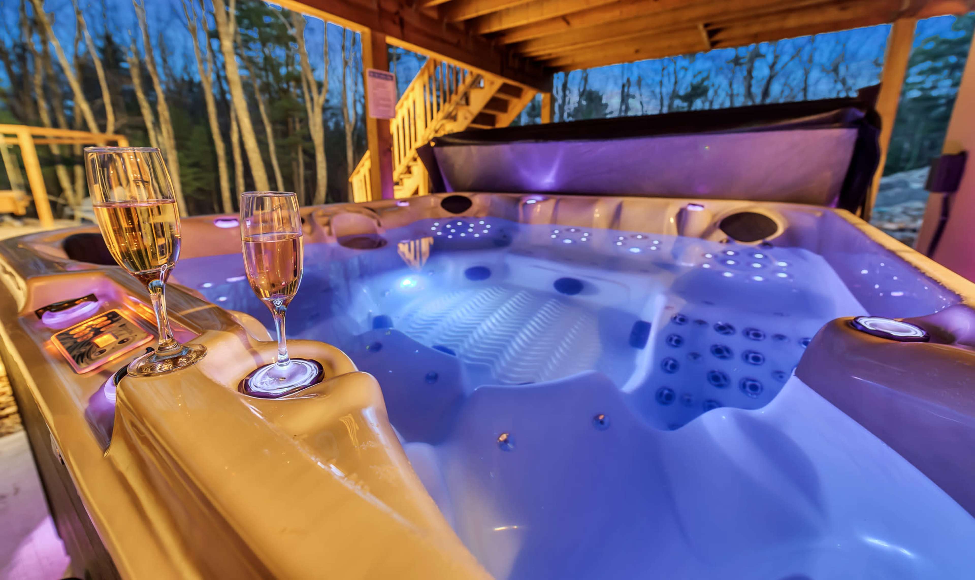 A hot tub under a wooden gazebo, with two champagne glasses placed on its edge and colorful lights illuminating the water.