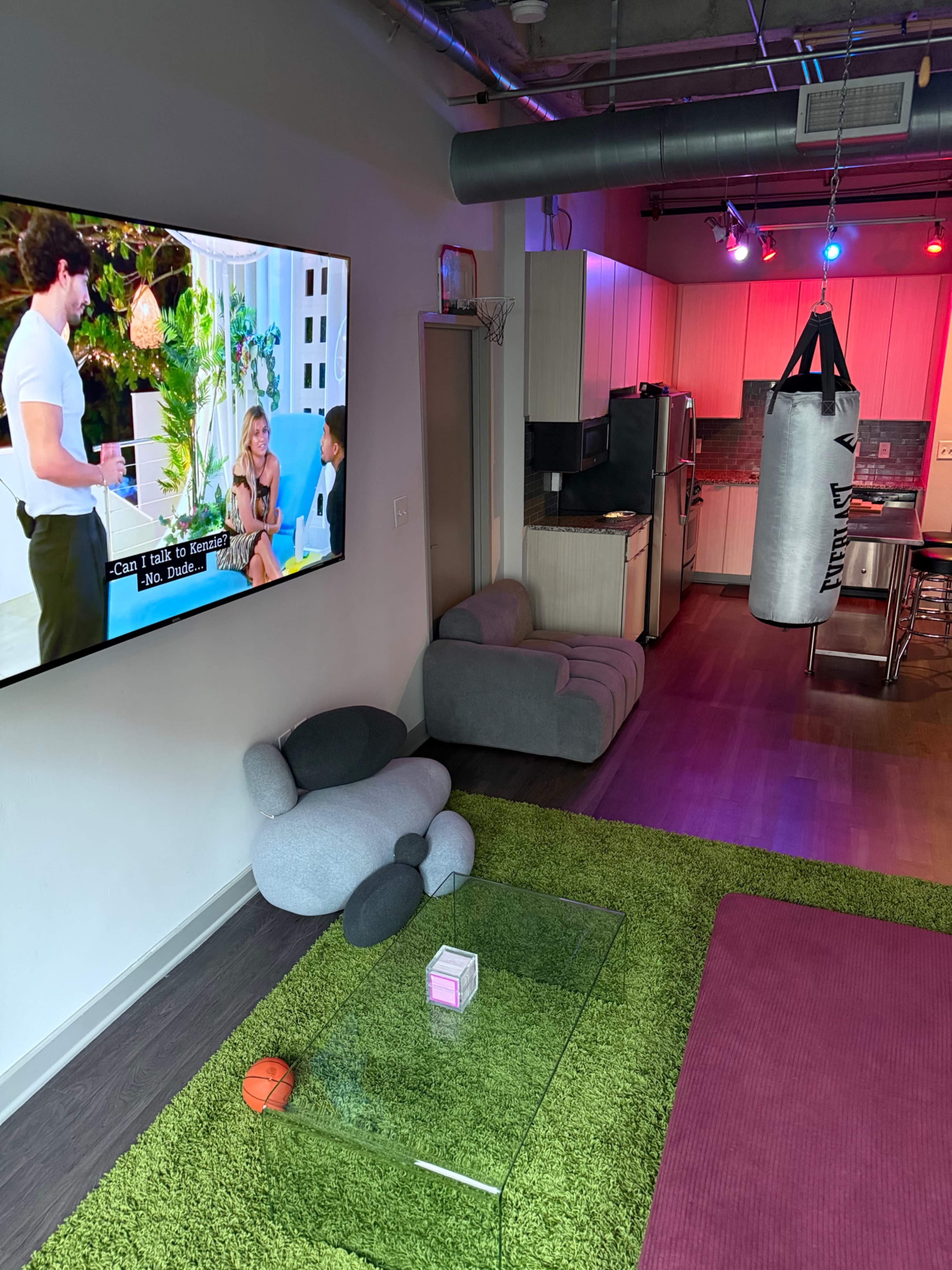 A cozy living space features a large television displaying a program, a green rug, a hanging punching bag, and a kitchen area in the background.