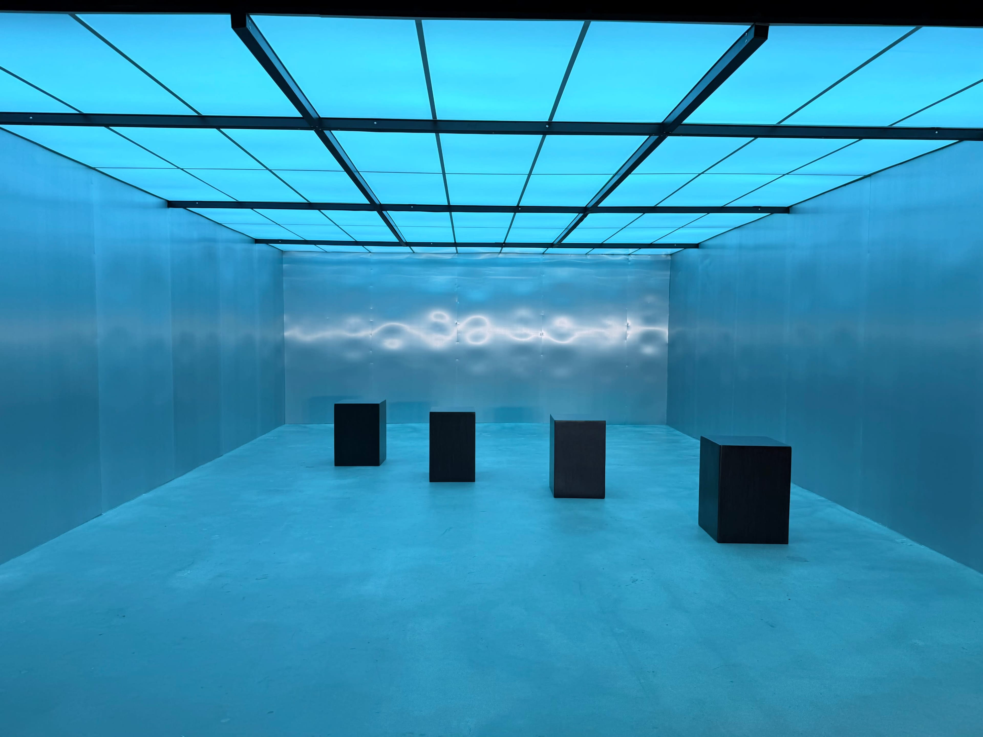 The image shows a minimalist exhibition space with four dark rectangular blocks set on a light-colored floor, illuminated by a blue-tinted ceiling.