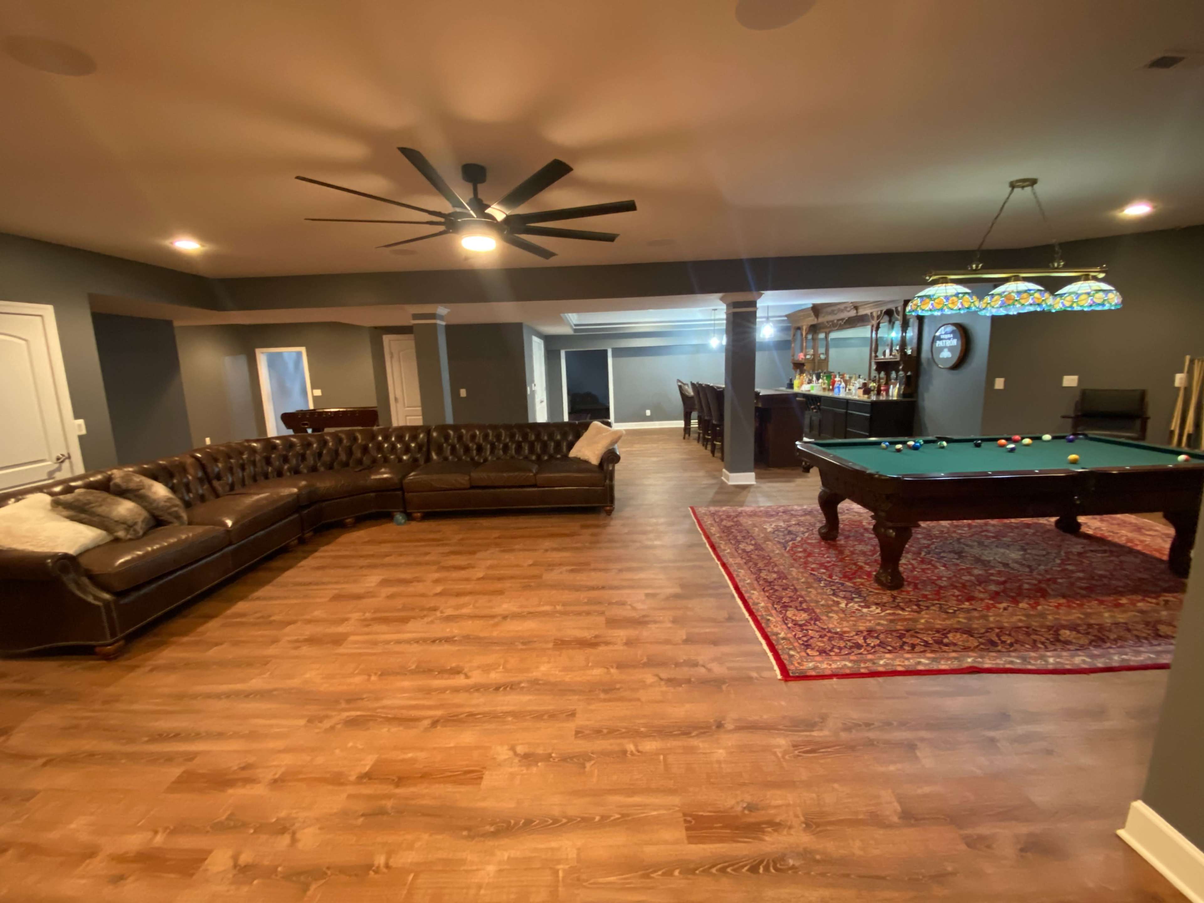 A basement recreation room featuring a pool table, a vintage-style sofa, and a bar with high stools in the background.