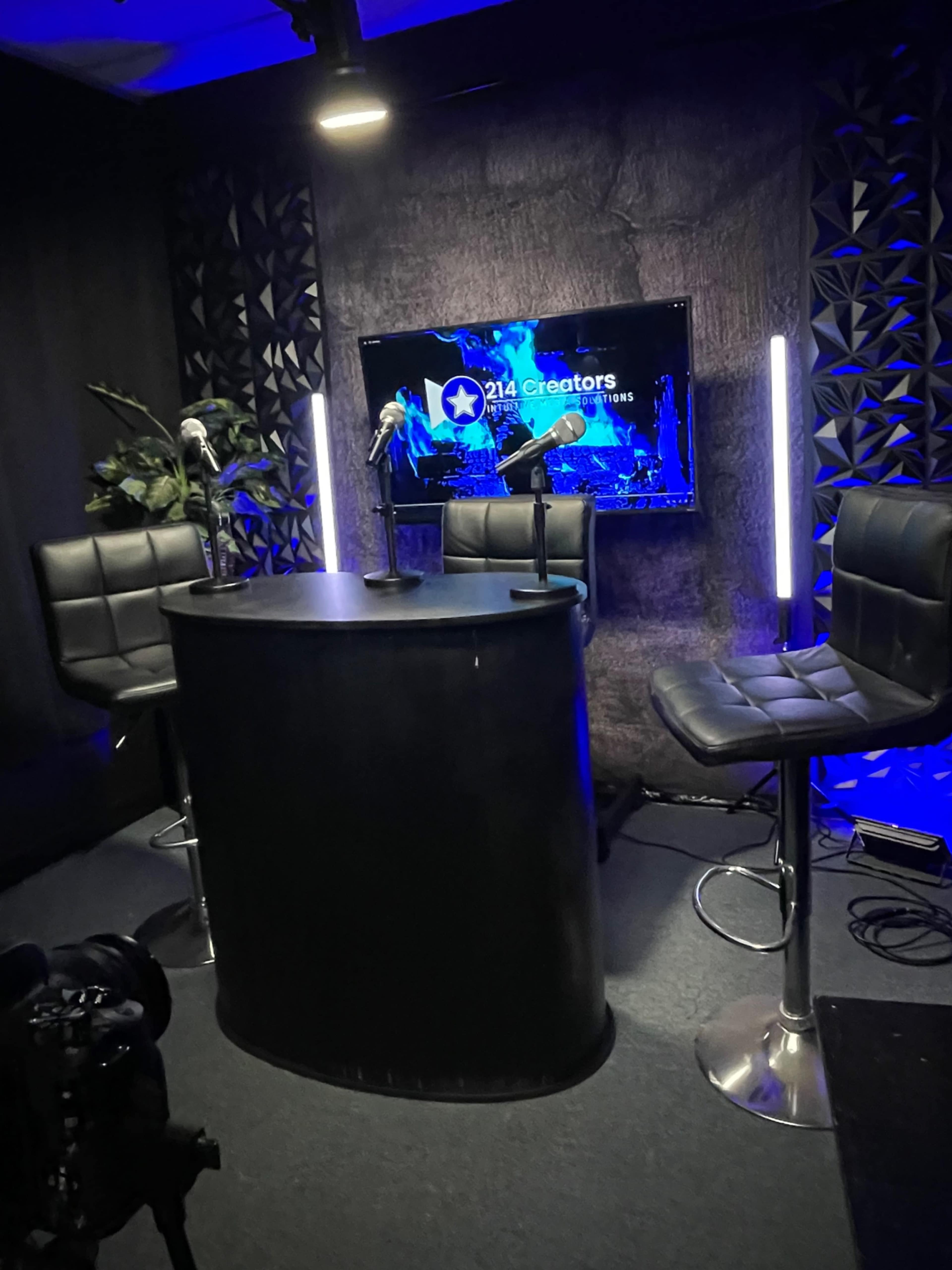 A modern studio setup with a round black table, three microphones, and high-backed bar stools, illuminated by blue lights and a screen displaying "214 Creators."