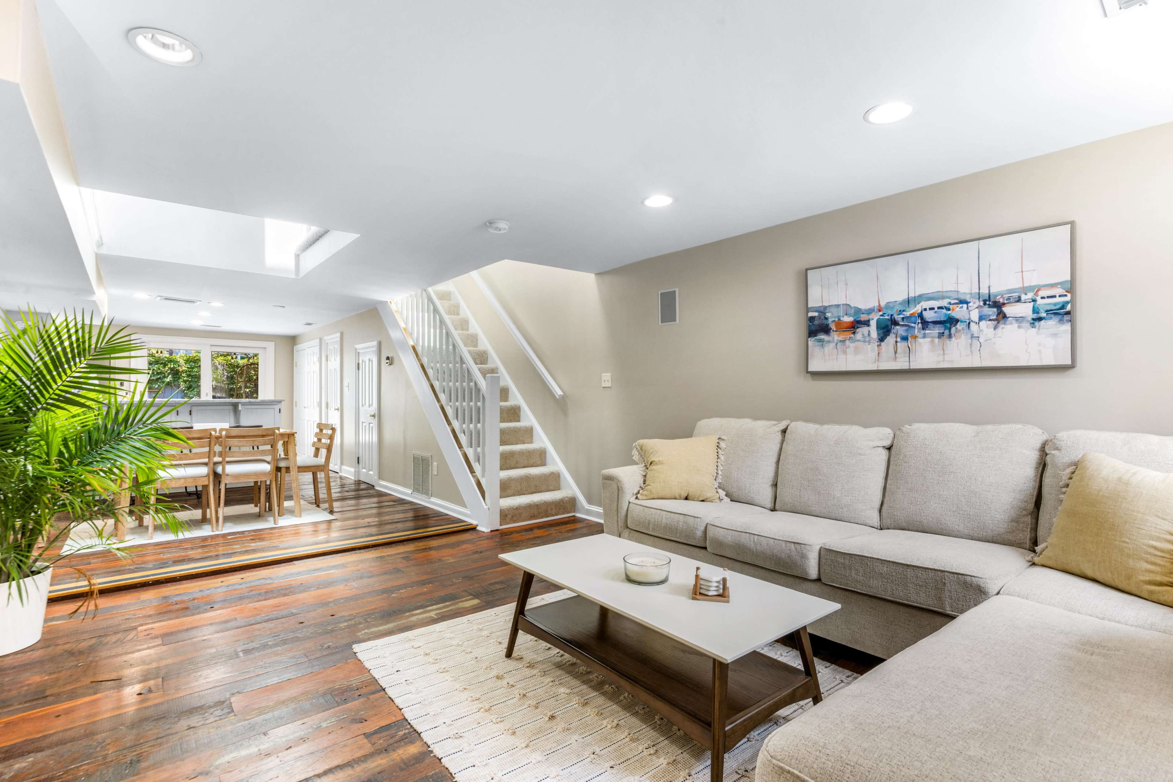 A cozy living area features a light-colored sectional sofa and a coffee table, with a staircase leading to a dining area illuminated by natural light.