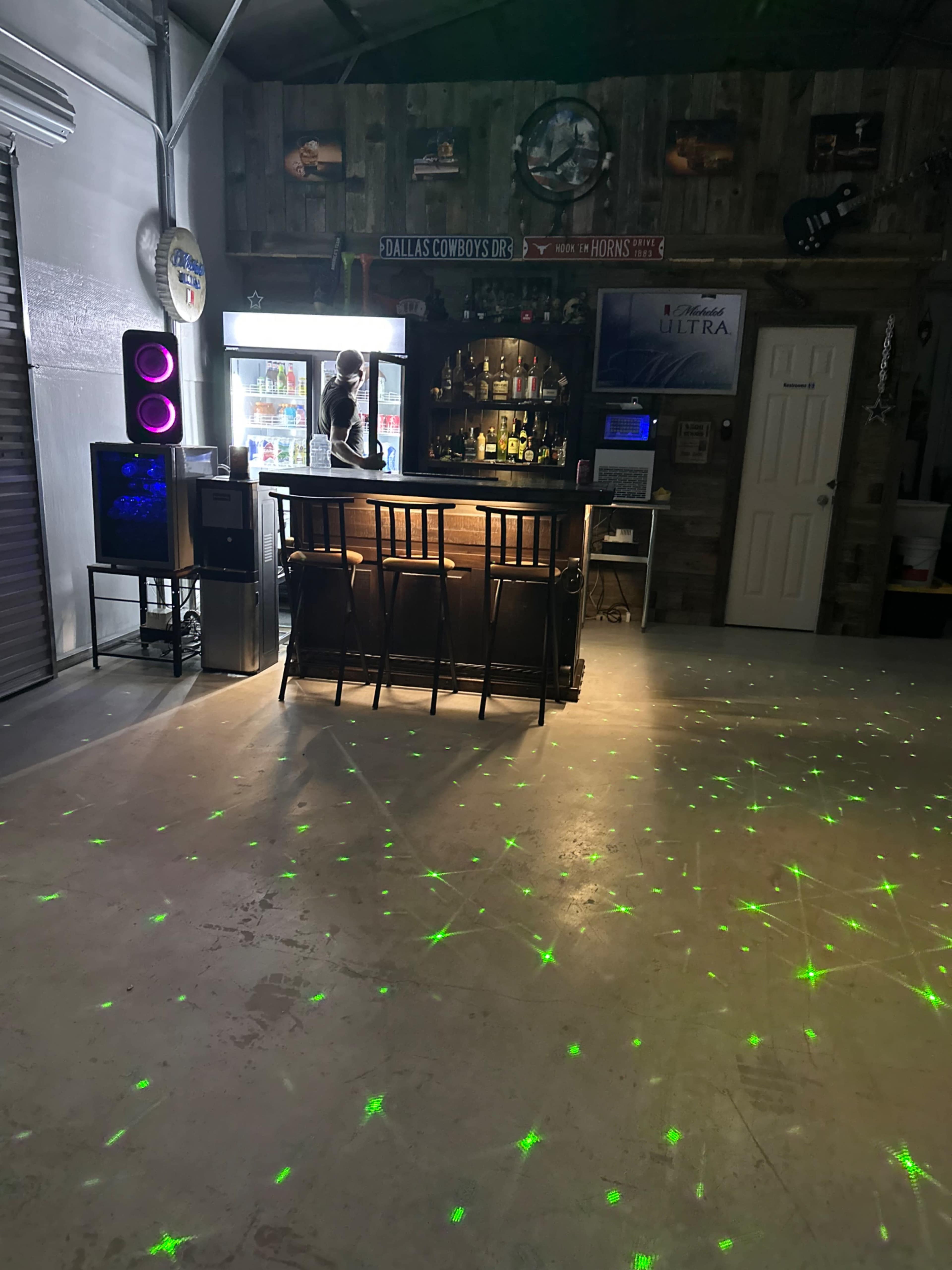 The image shows a bar area inside a garage with a counter, chairs, and a cooler, while green laser lights cover the floor.