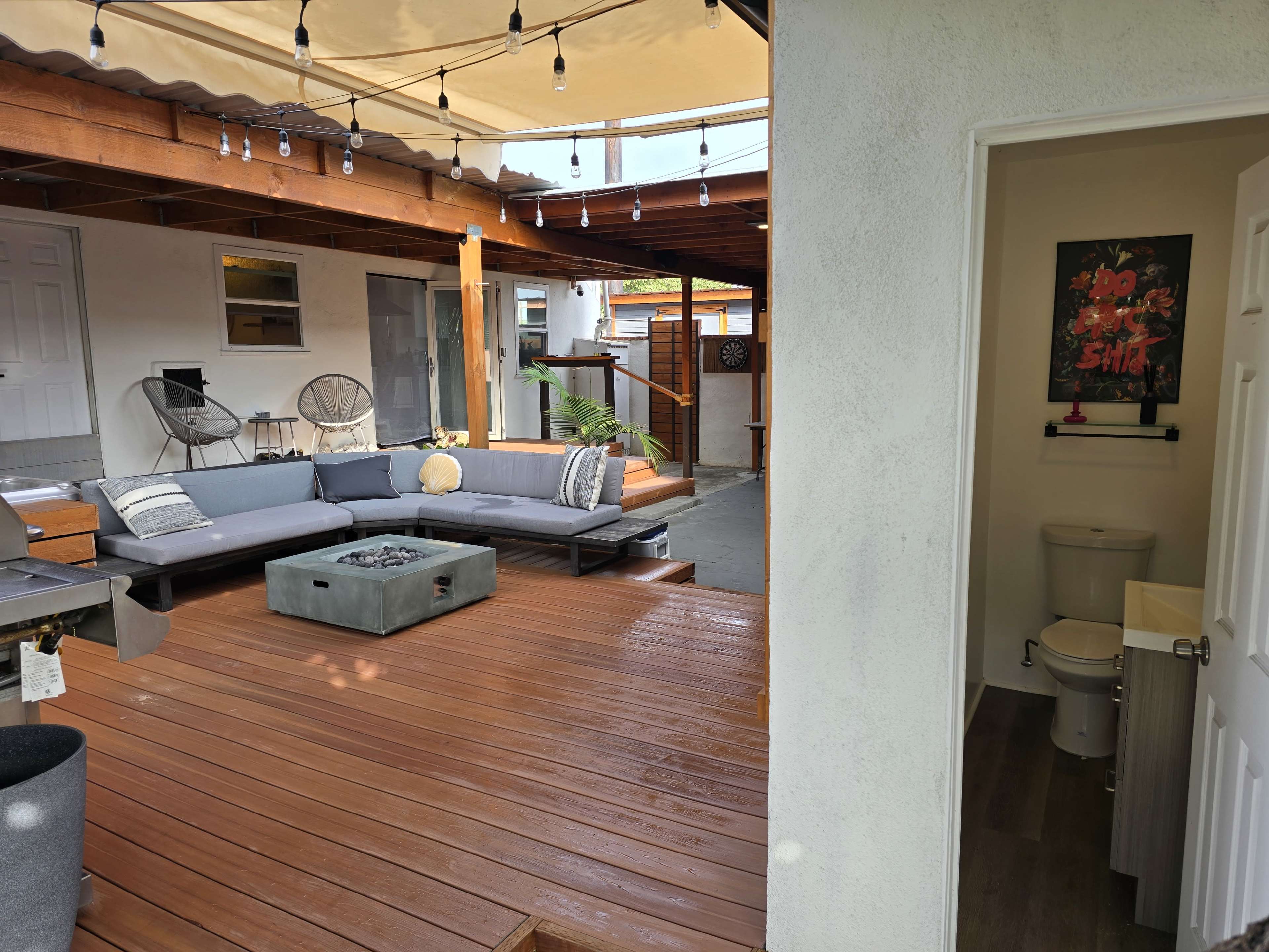 The image shows a modern outdoor living space featuring a wooden deck with a large sectional sofa, a fire pit in the center, and a nearby bathroom entrance.