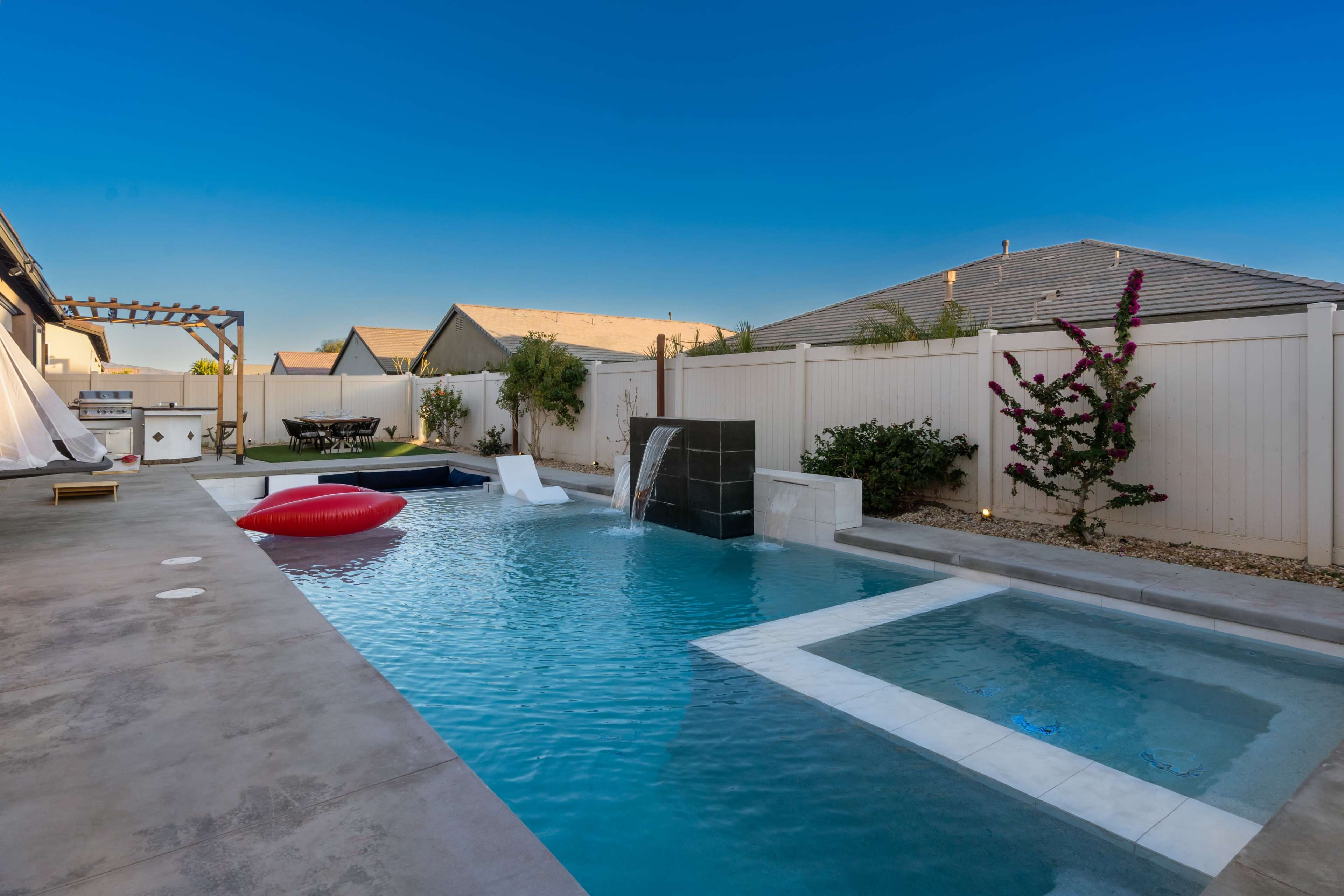 A modern backyard with a swimming pool, a wall fountain, and a seating area surrounded by houses and landscaping.