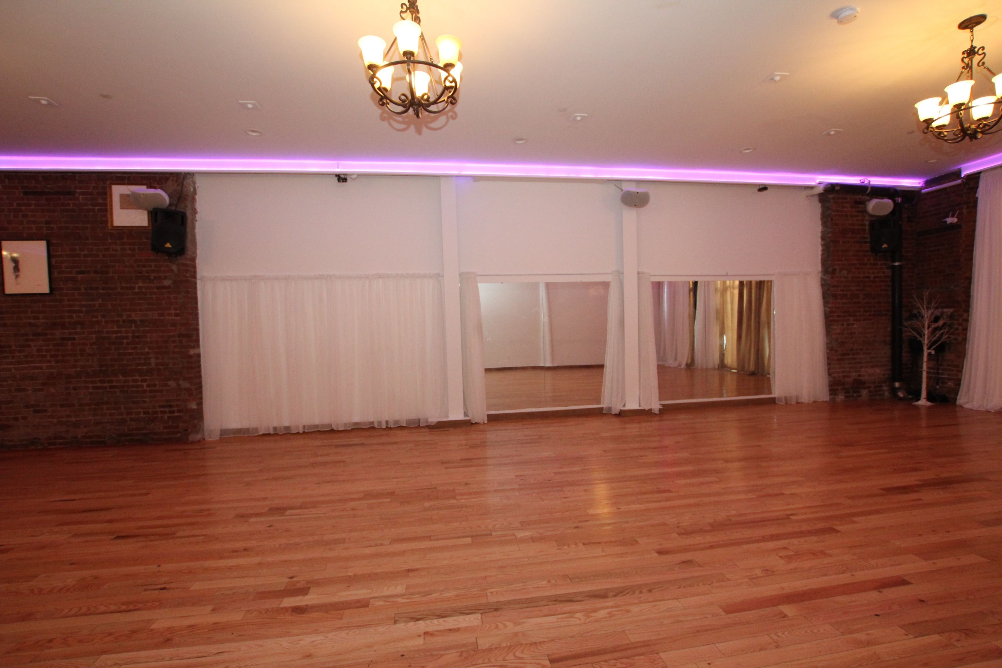 A spacious room with hardwood floors, exposed brick walls, and large mirrors, illuminated by chandeliers and soft purple lighting.