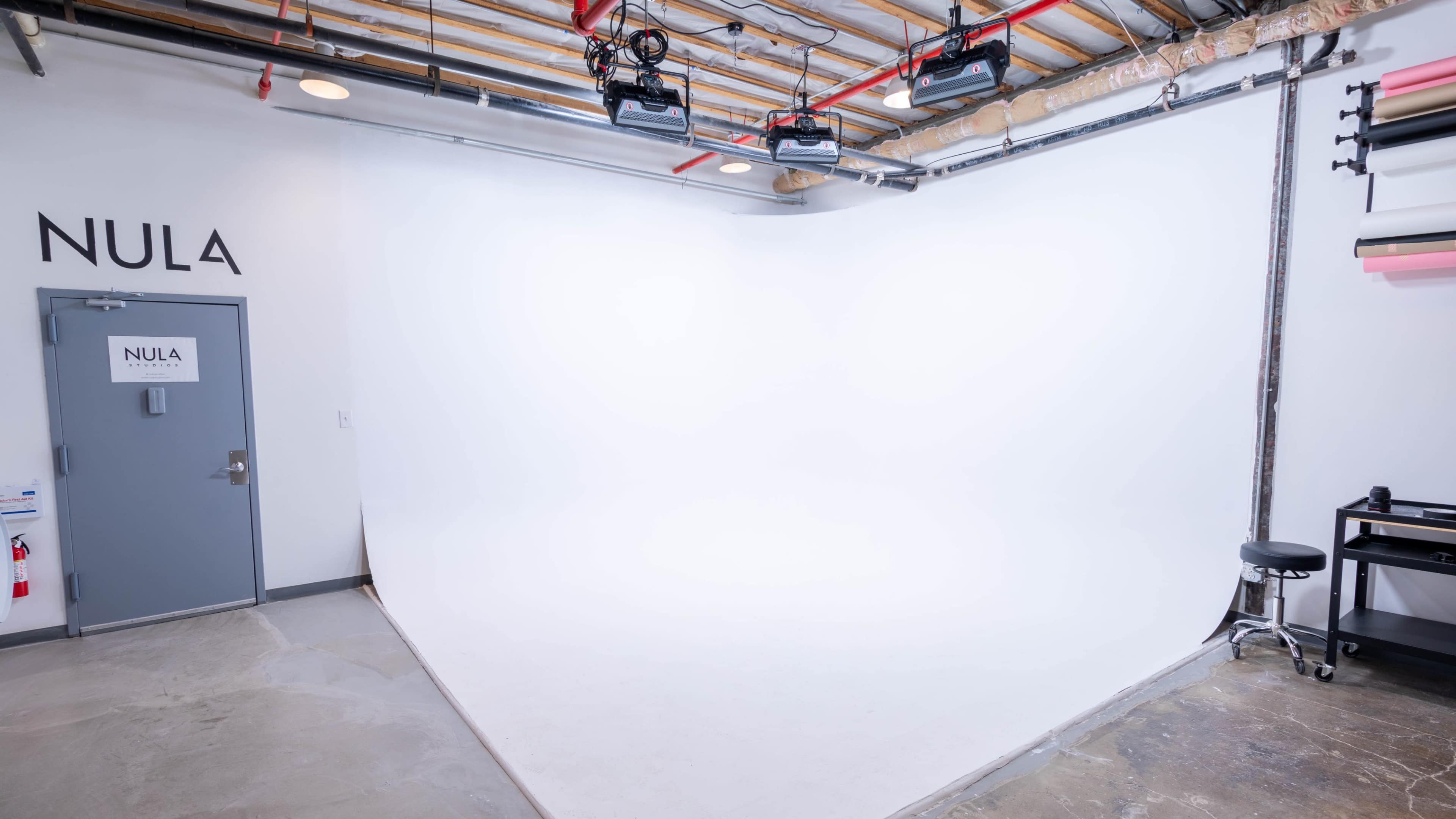 A photography studio with a white backdrop, a gray door labeled "NULA," and several lights mounted on the ceiling.