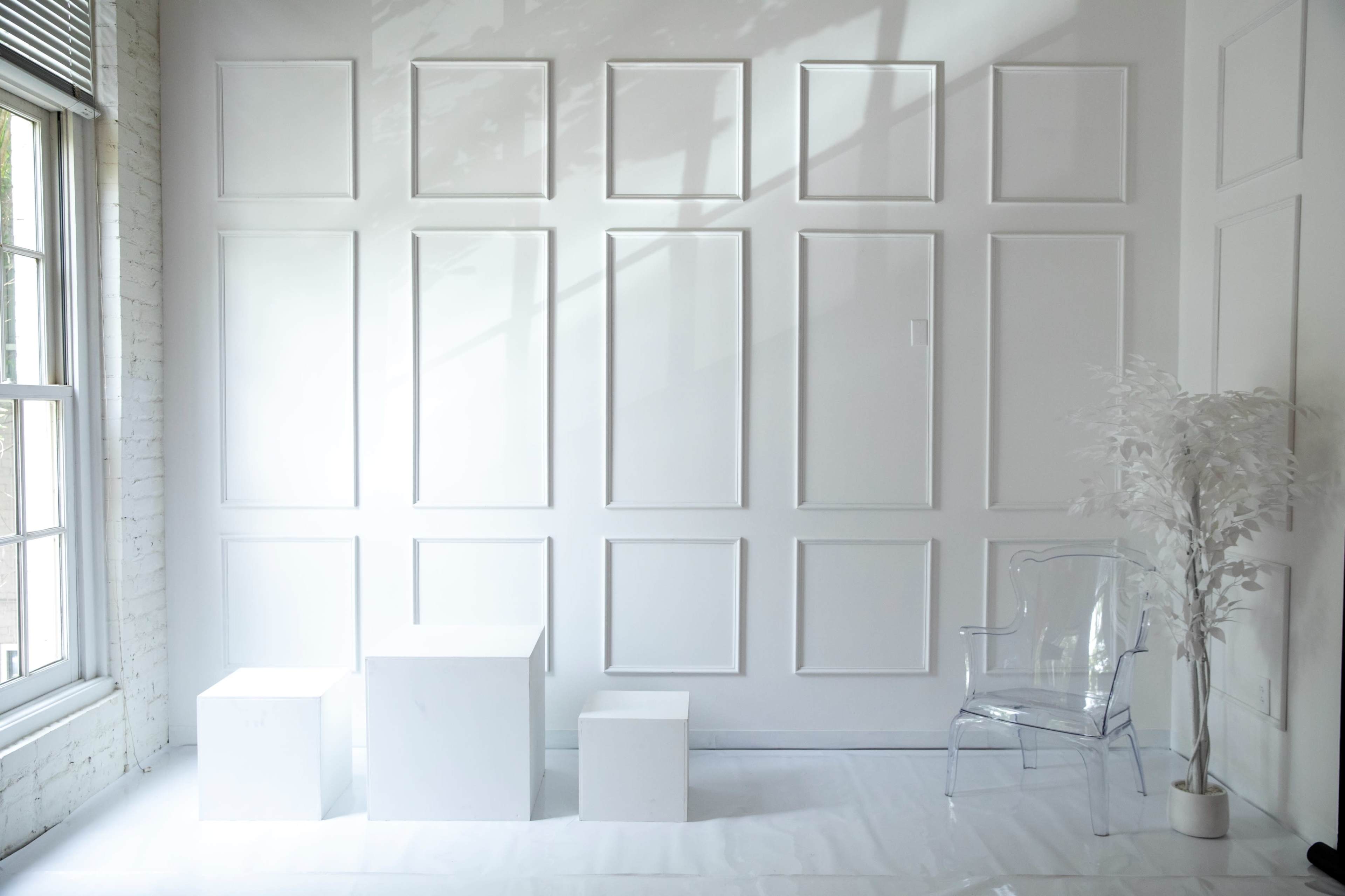 The image shows a minimalistic interior space with a white wall featuring rectangular panels, three white geometric shapes on the floor, and a clear acrylic chair beside a decorative white plant.