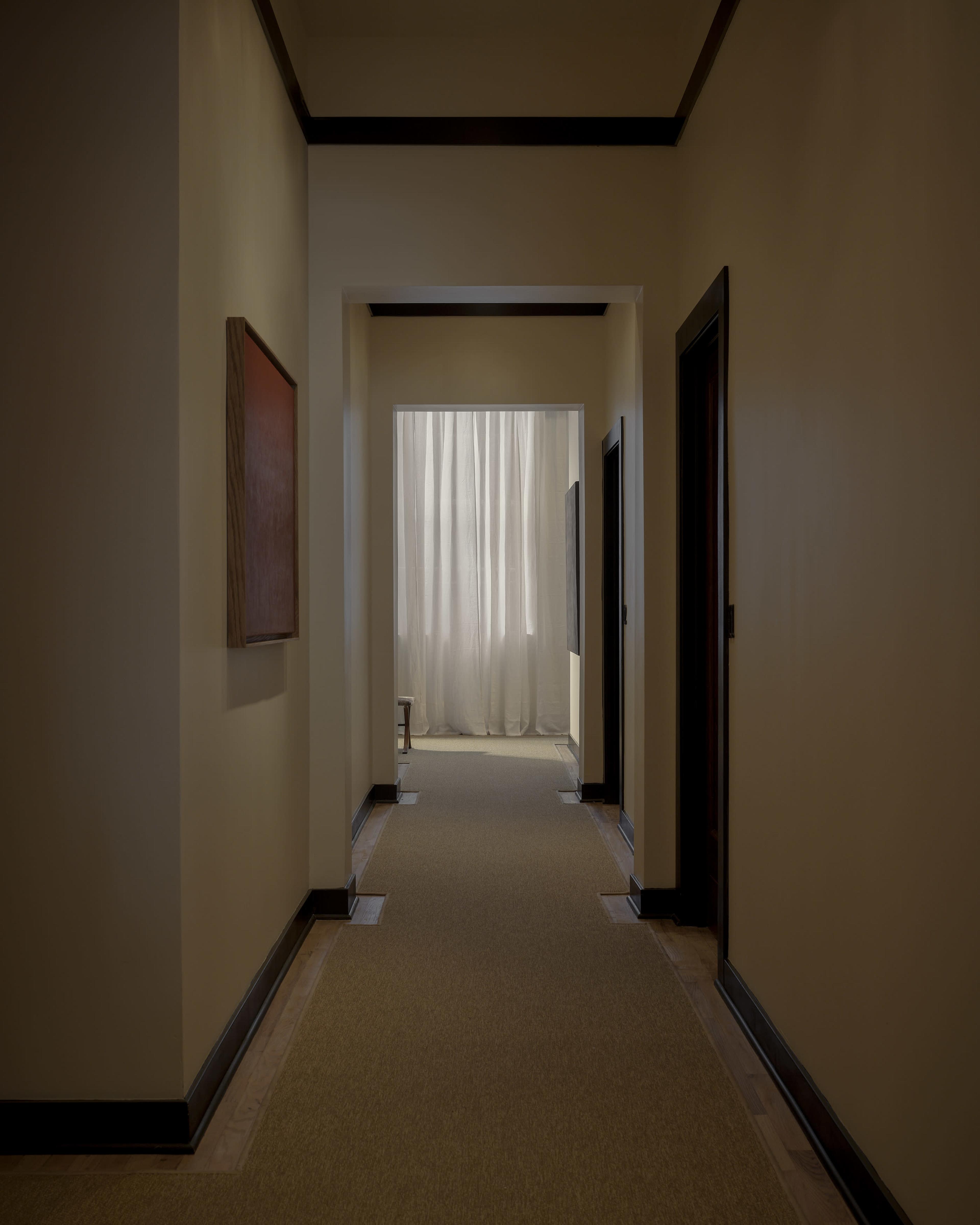 A narrow hallway with beige walls and a patterned carpet leads to a bright area partially obscured by white curtains.