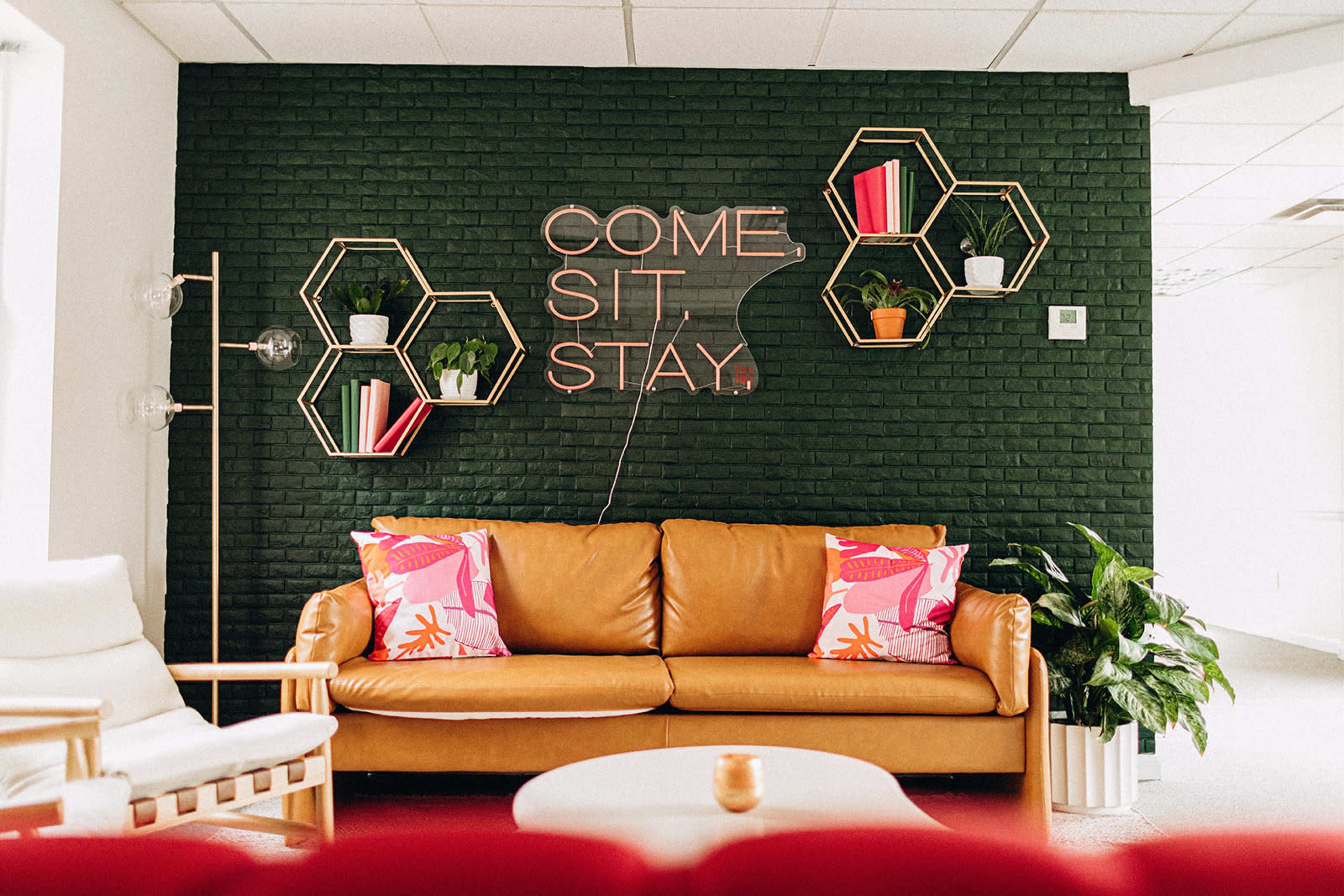 A brown leather couch is positioned against a dark green brick wall, adorned with a neon sign and hexagonal shelves featuring plants and decorative books.