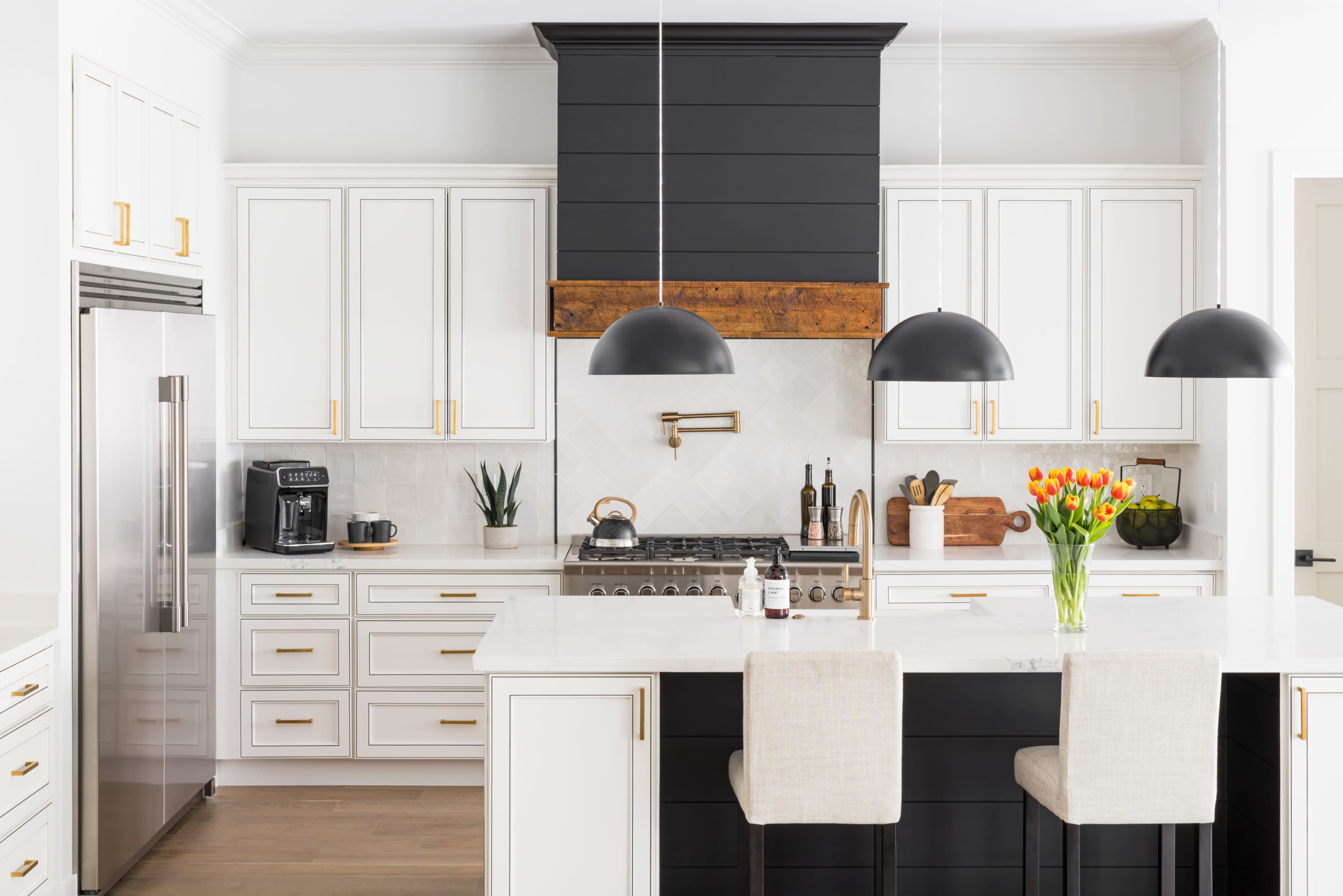 A modern kitchen features white cabinetry, a black hood over the stove, and three pendant lights, with a central island and bar stools.