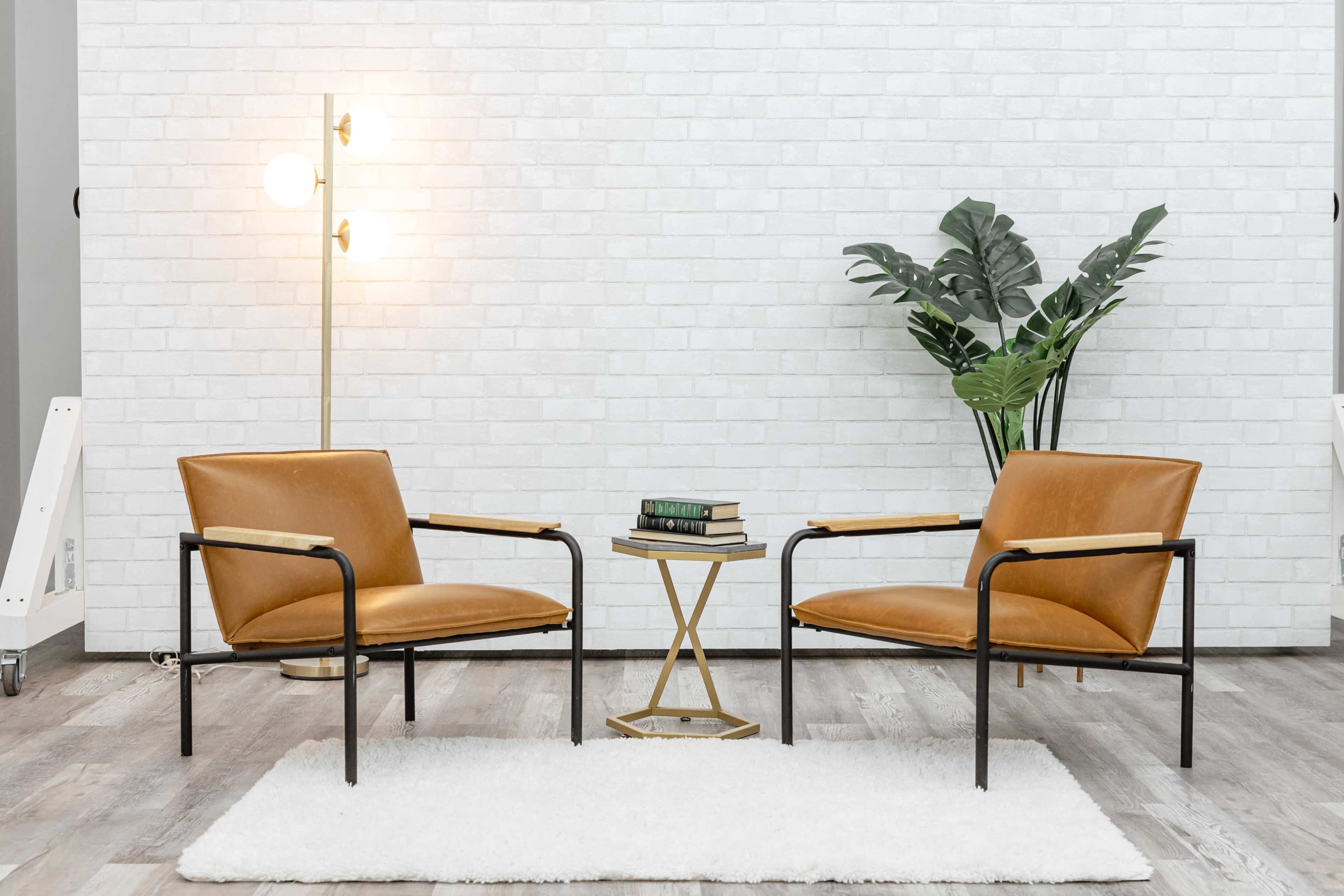 Two brown chairs are positioned opposite each other with a small gold side table and a stack of books in between, set against a white brick wall and a potted plant in the background.