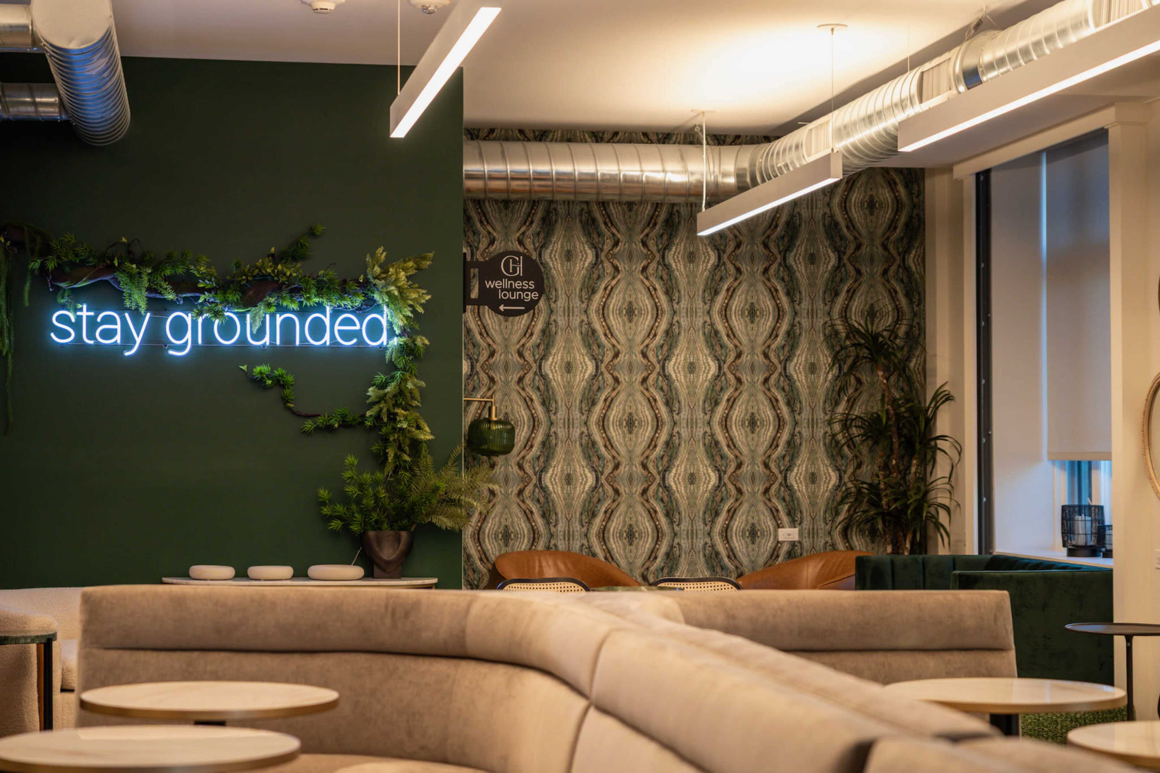 The image shows a modern lounge area with green walls, a neon sign that says "stay grounded," and stylish seating arrangements.