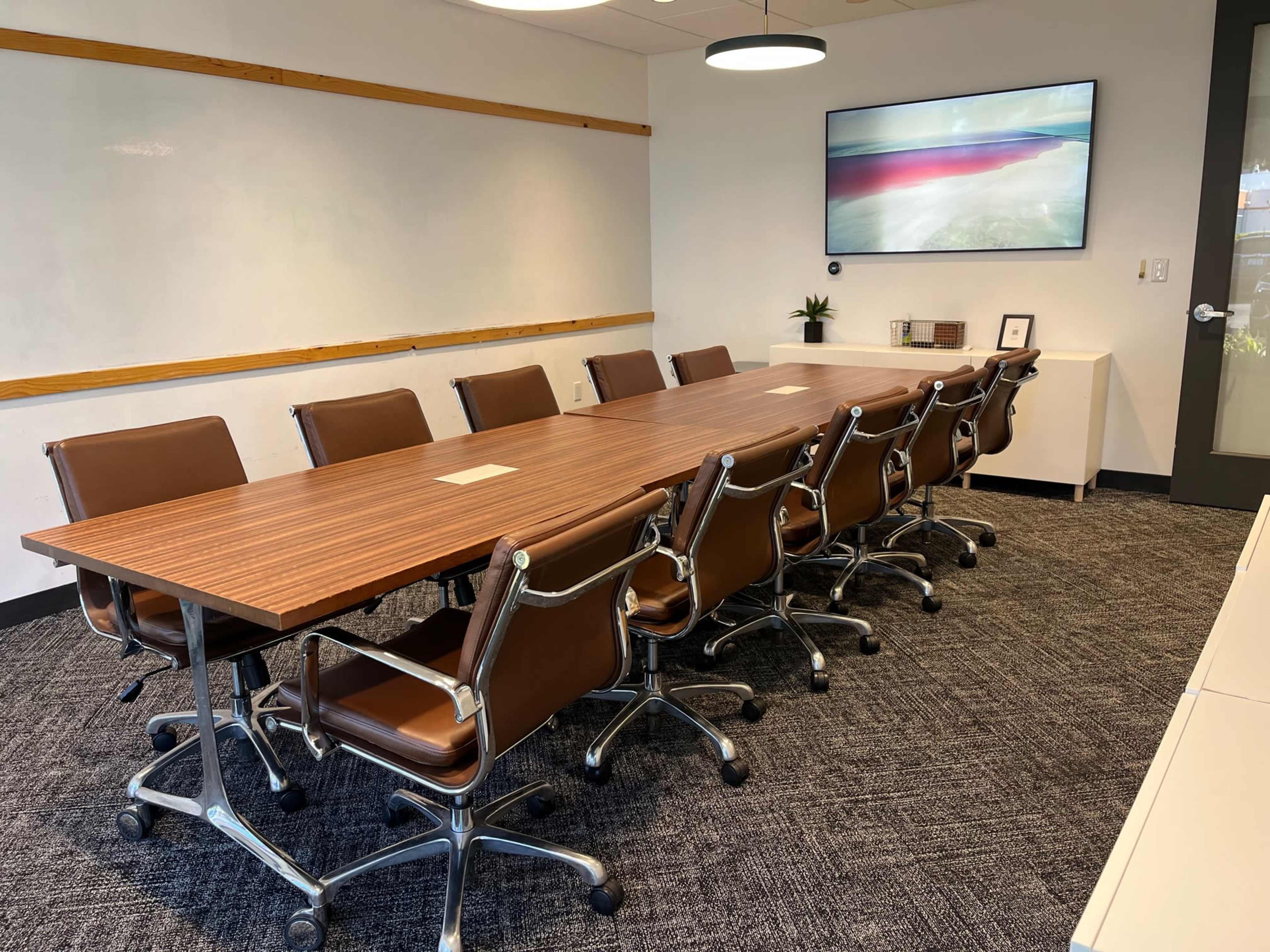 A conference room features a long wooden table surrounded by eight brown leather chairs and a wall-mounted screen displaying an abstract image.
