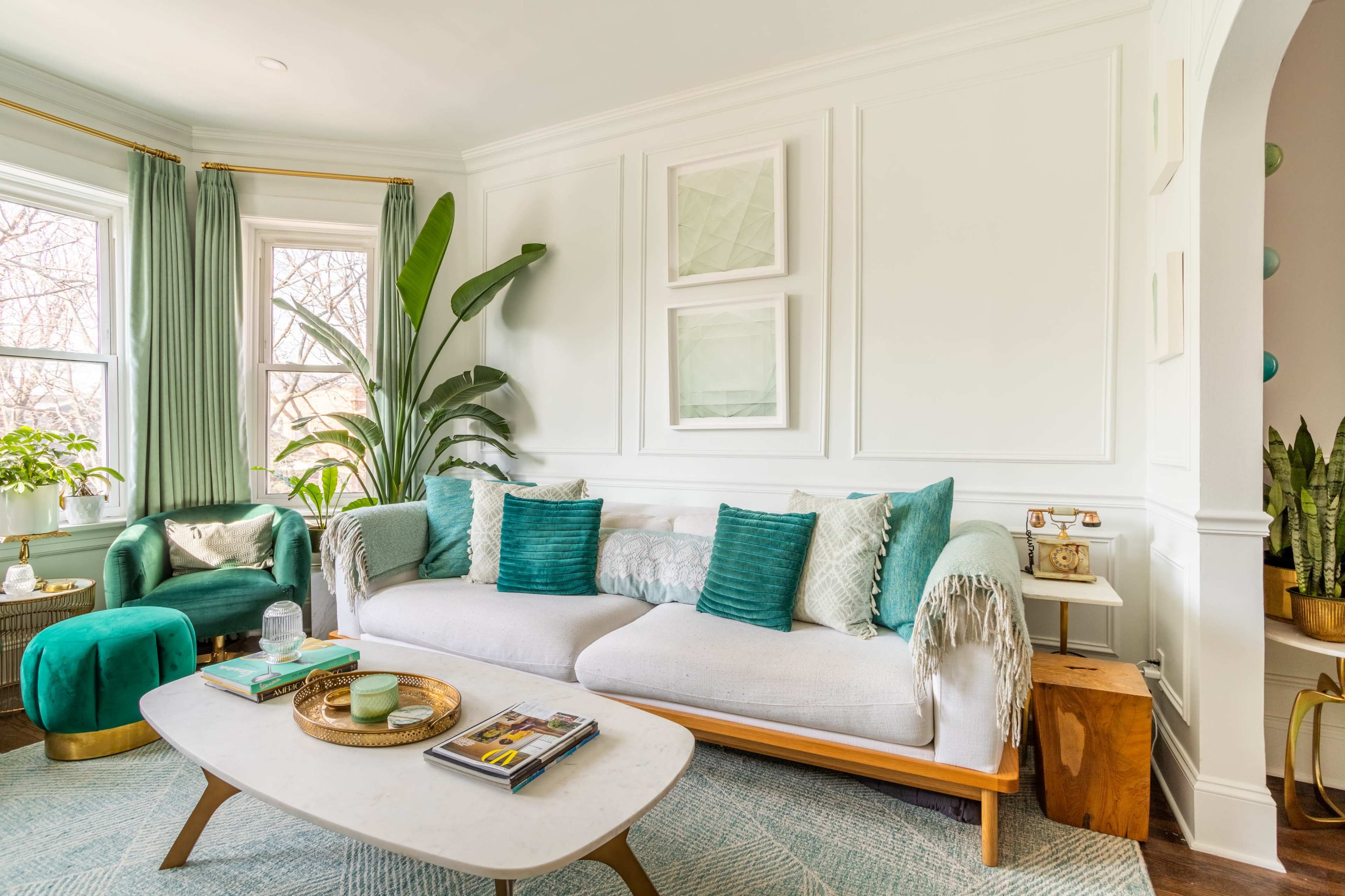 A living room features a light gray sofa with teal cushions, a round green ottoman, large windows with green curtains, and various houseplants.