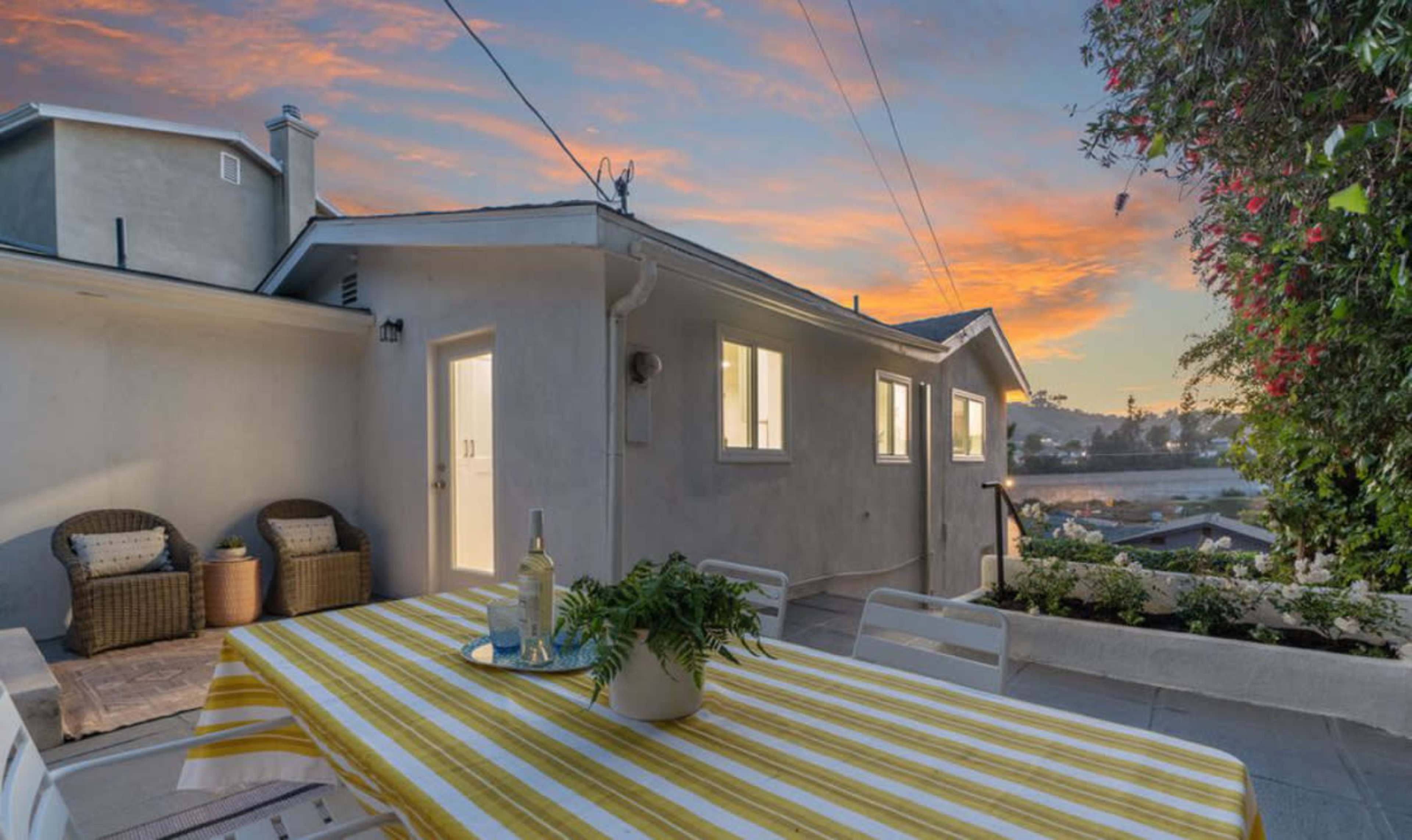 The image shows a patio with a striped tablecloth, a potted plant, and two wicker chairs, alongside a house and a colorful sunset sky.