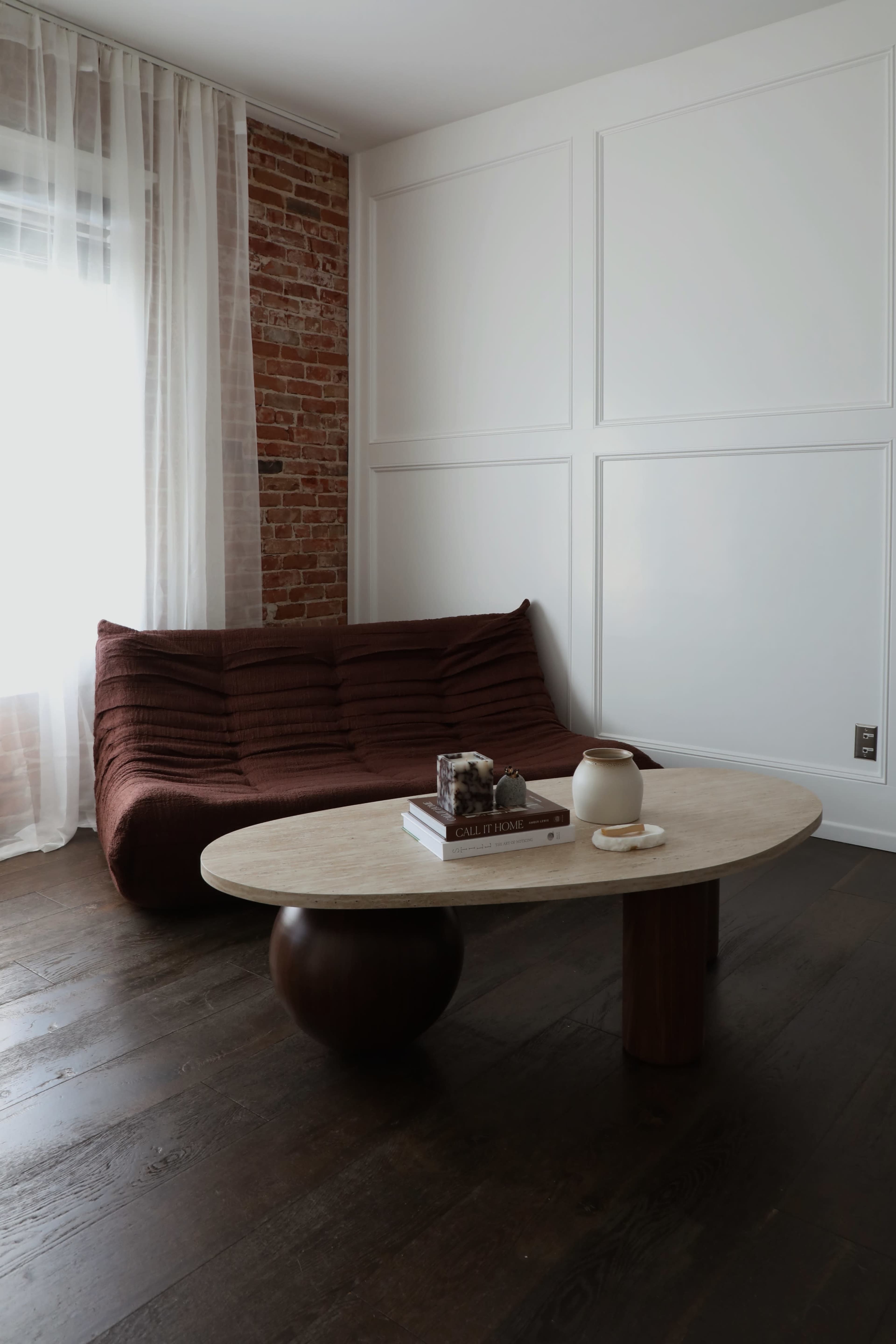 The image shows a minimalist living room featuring a maroon sofa, a round beige coffee table, and a textured wall with exposed brick.
