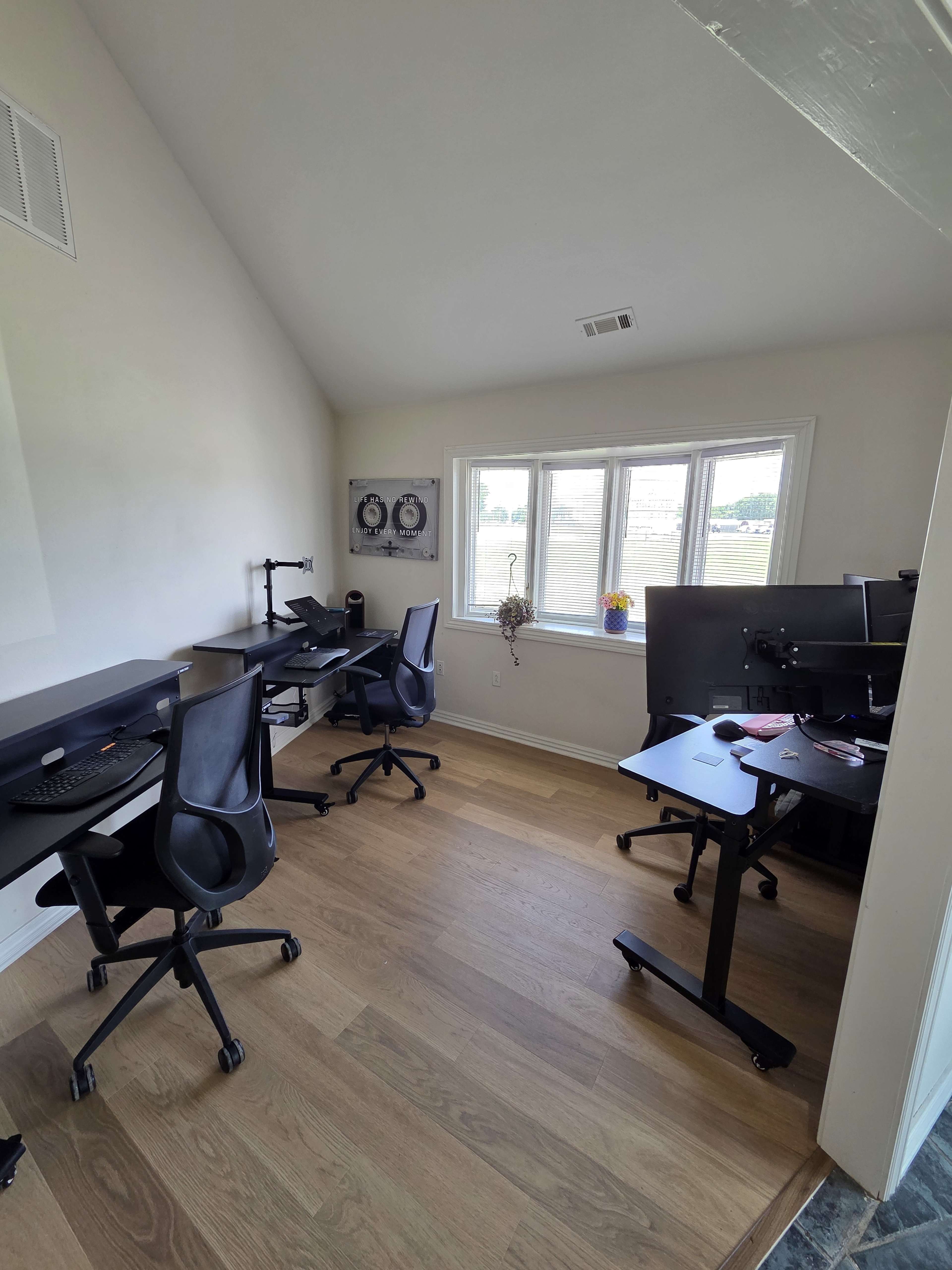 A sunlit home office features four black office chairs and desks arranged against the walls, with large windows offering a view outside.