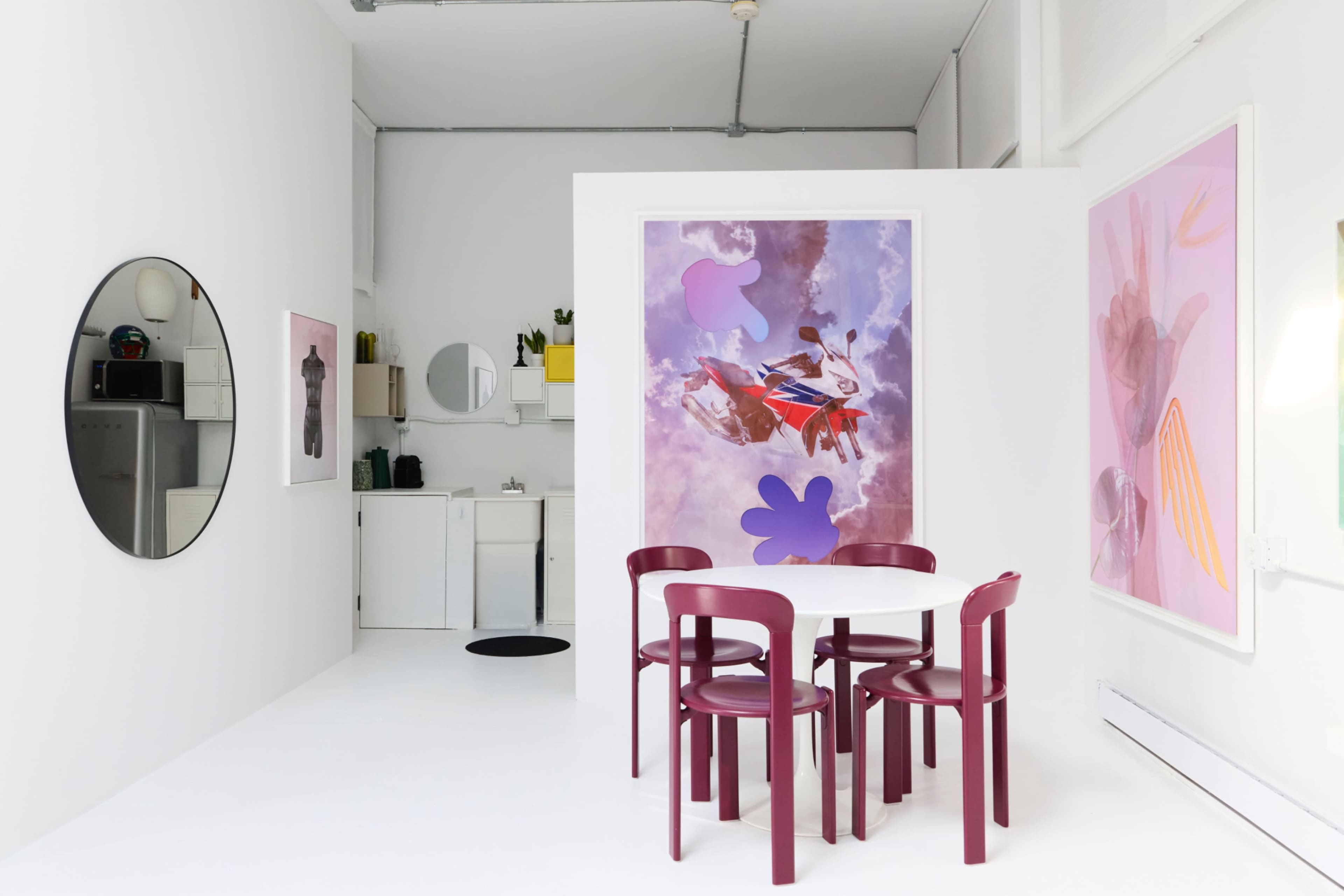 The image shows a modern, minimalist room with a round table and four red chairs, a kitchenette area in the background, and colorful artwork on the walls.