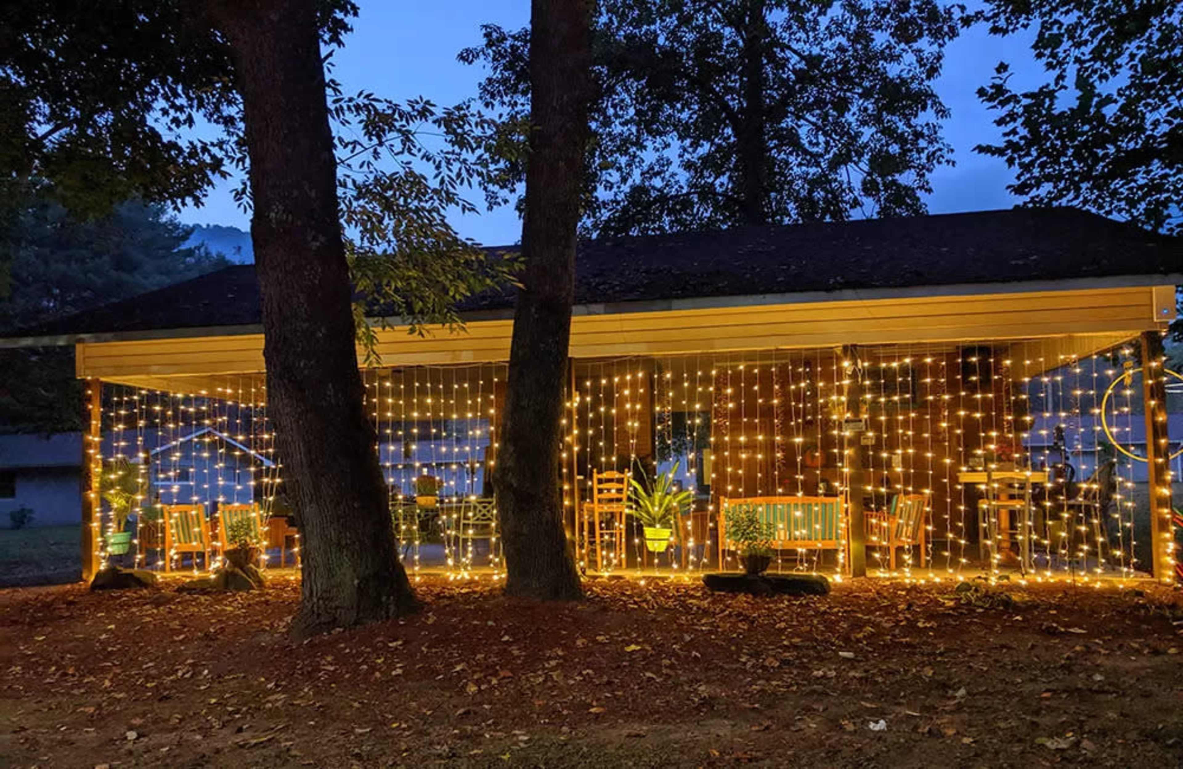 A porch illuminated by decorative string lights, surrounded by trees and a few chairs.