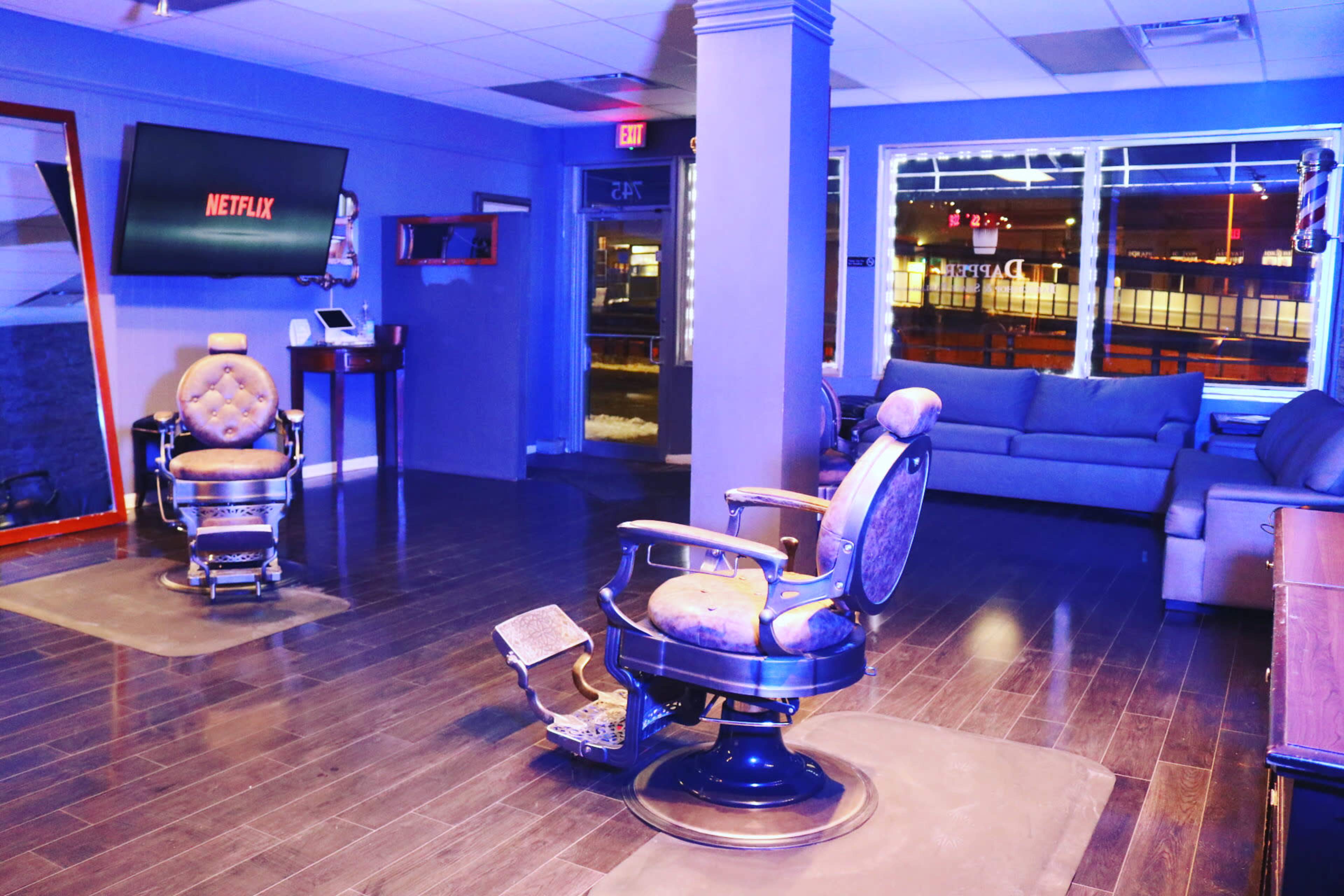 A modern barbershop interior featuring two barber chairs, a large screen displaying Netflix, and a seating area with a couch.