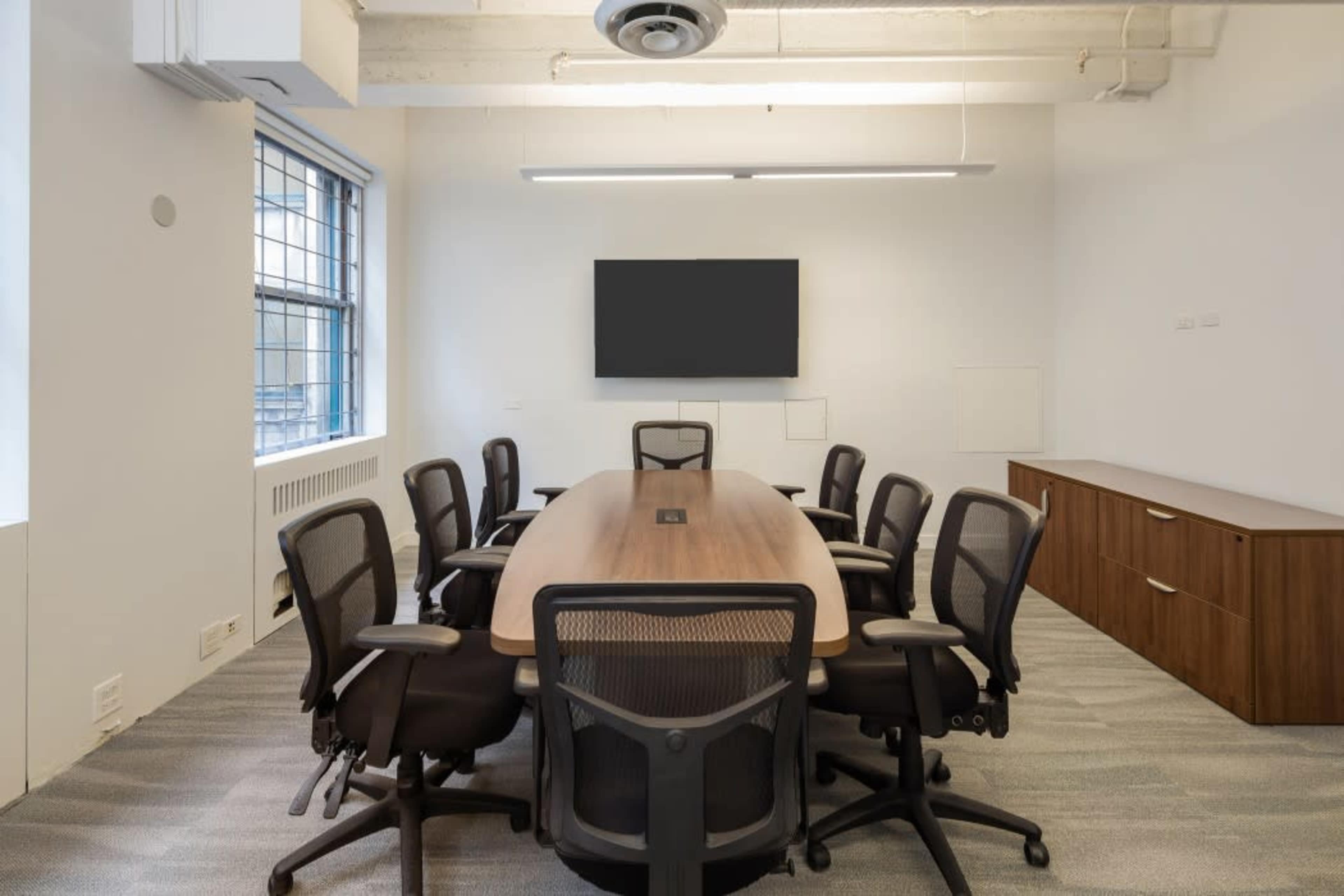 A conference room features a large wooden table surrounded by eight black office chairs, with a wall-mounted screen and windows providing natural light.