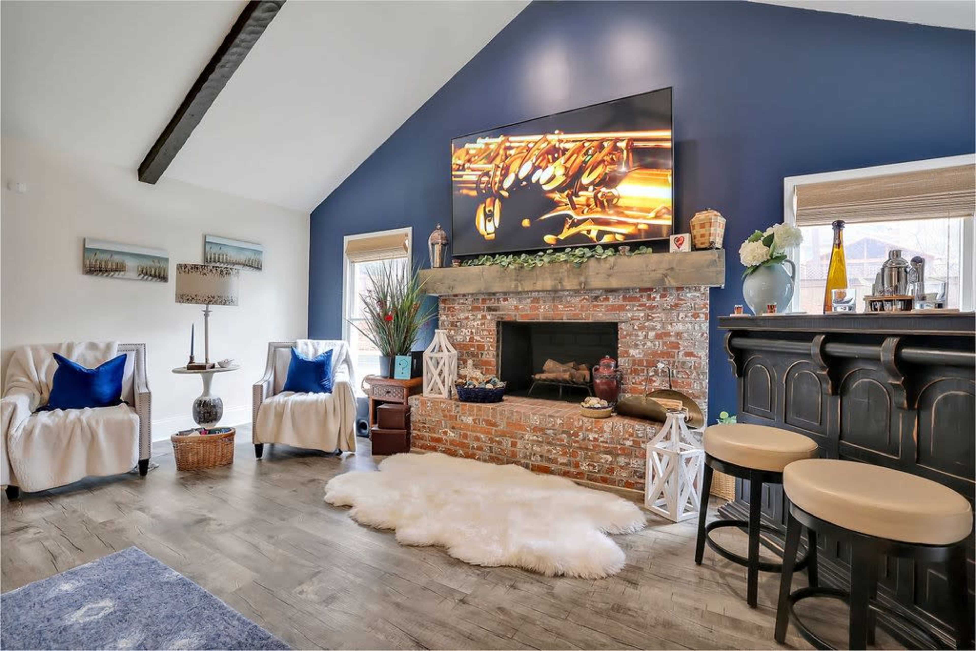 The room features a brick fireplace with a modern television above it, flanked by two armchairs, a bar area, and decorative elements on the walls.
