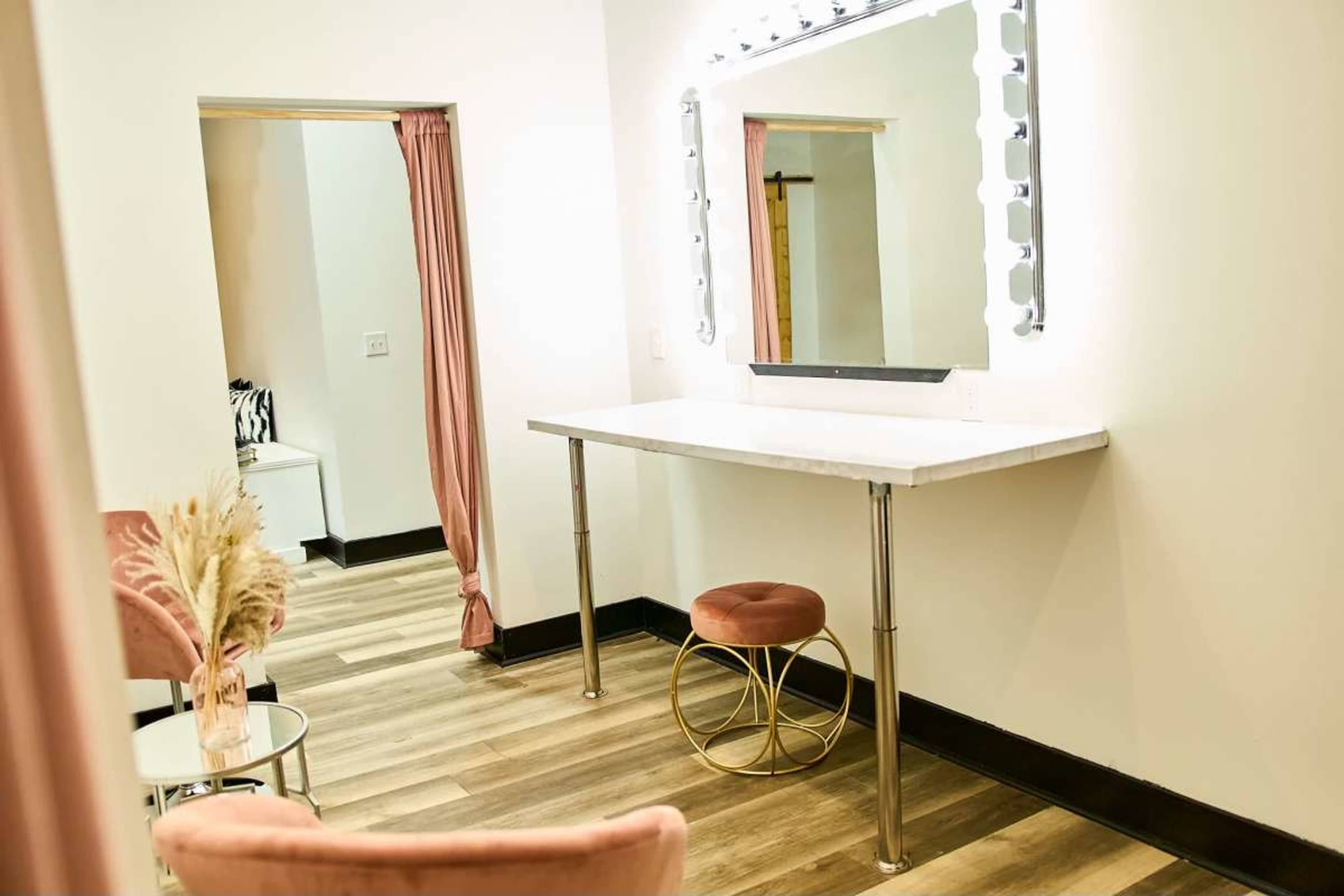 The image shows a minimally furnished vanity area with a large illuminated mirror, a marble-topped table, a circular stool, and pink curtains.