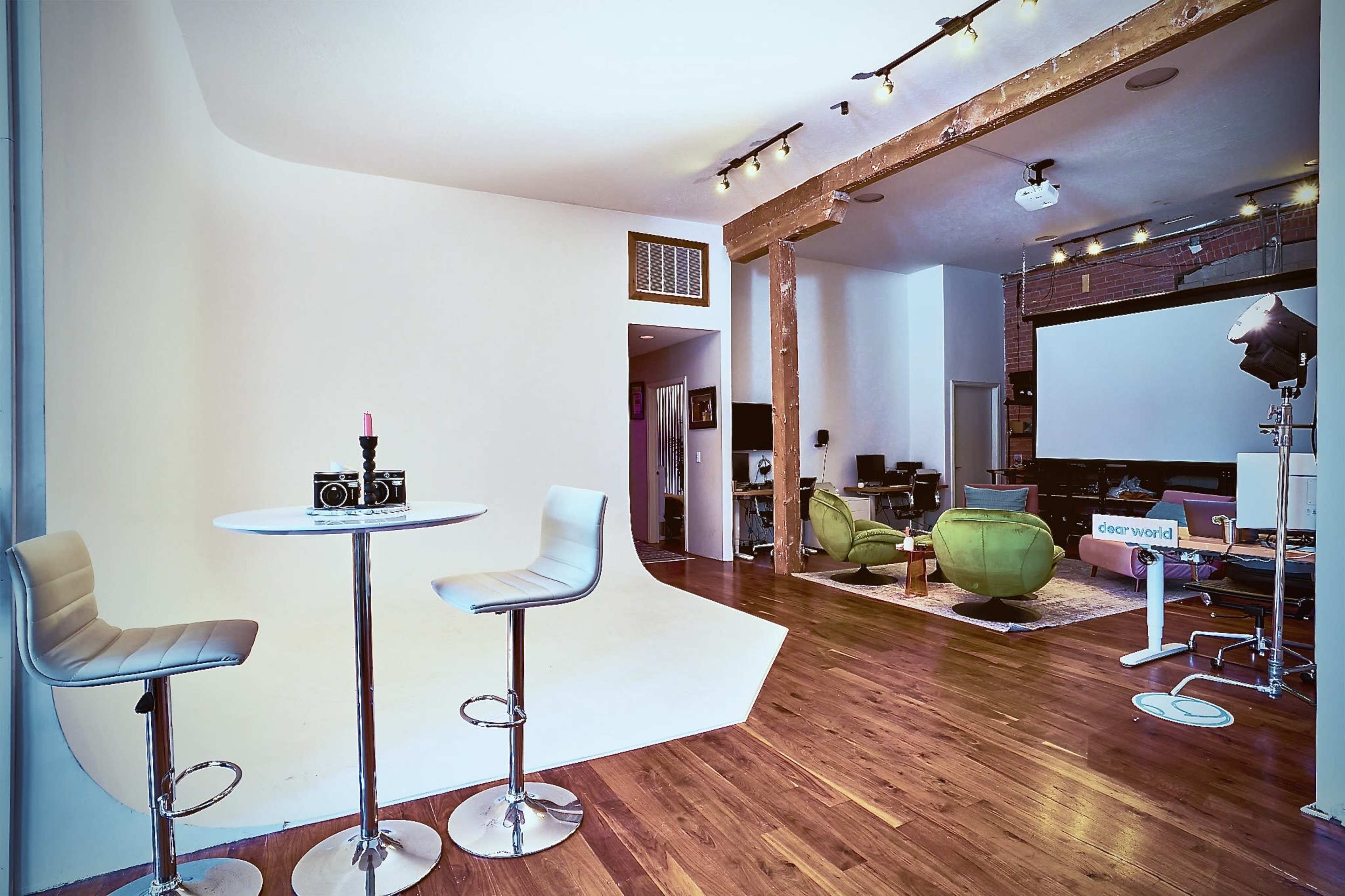 A modern studio space with a small round table and two stools in the foreground, hardwood floors, and various seating arrangements in the background.