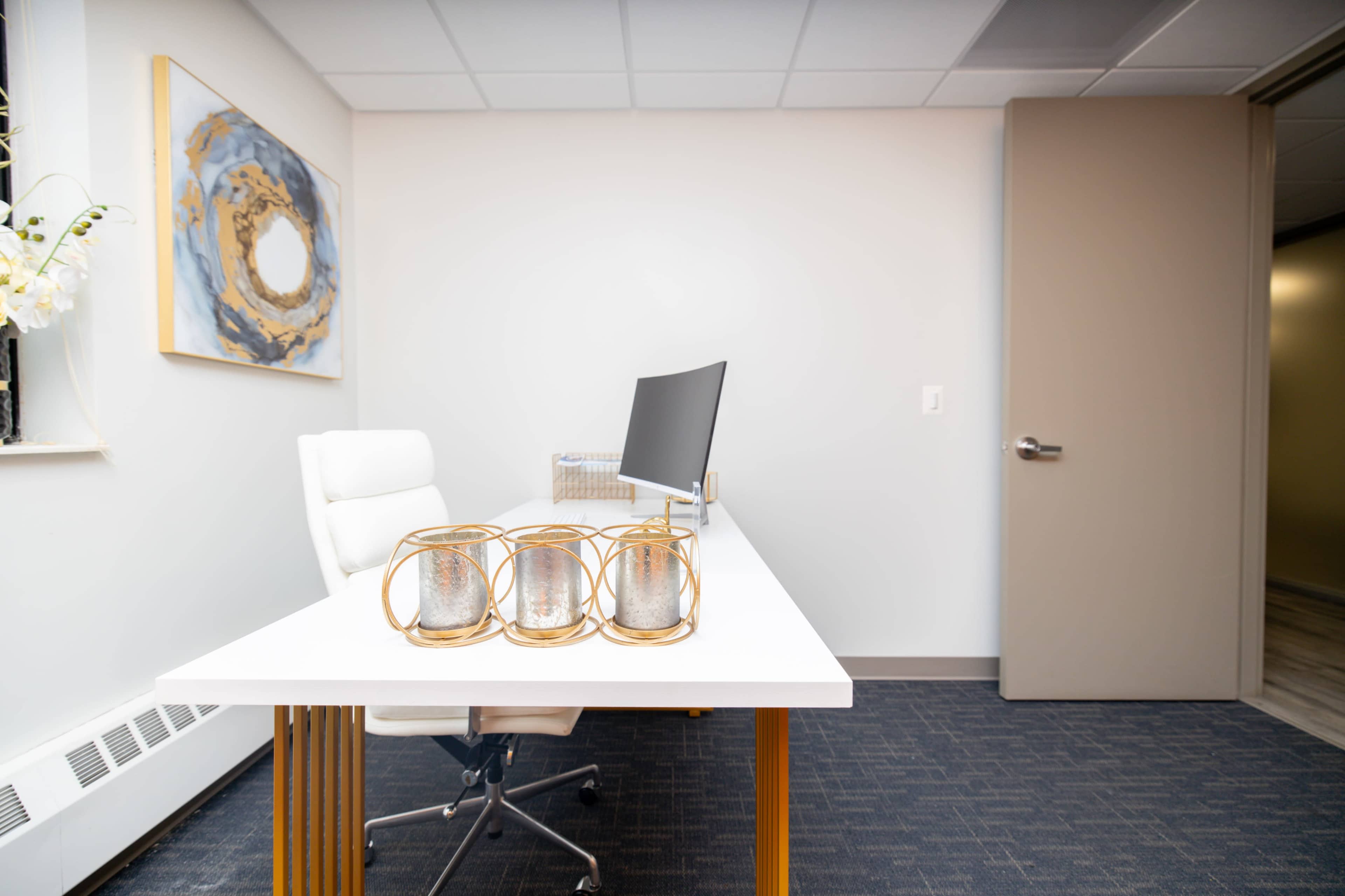 A modern office space features a desk with a computer, a white chair, and decorative containers, with a patterned artwork hanging on the wall.