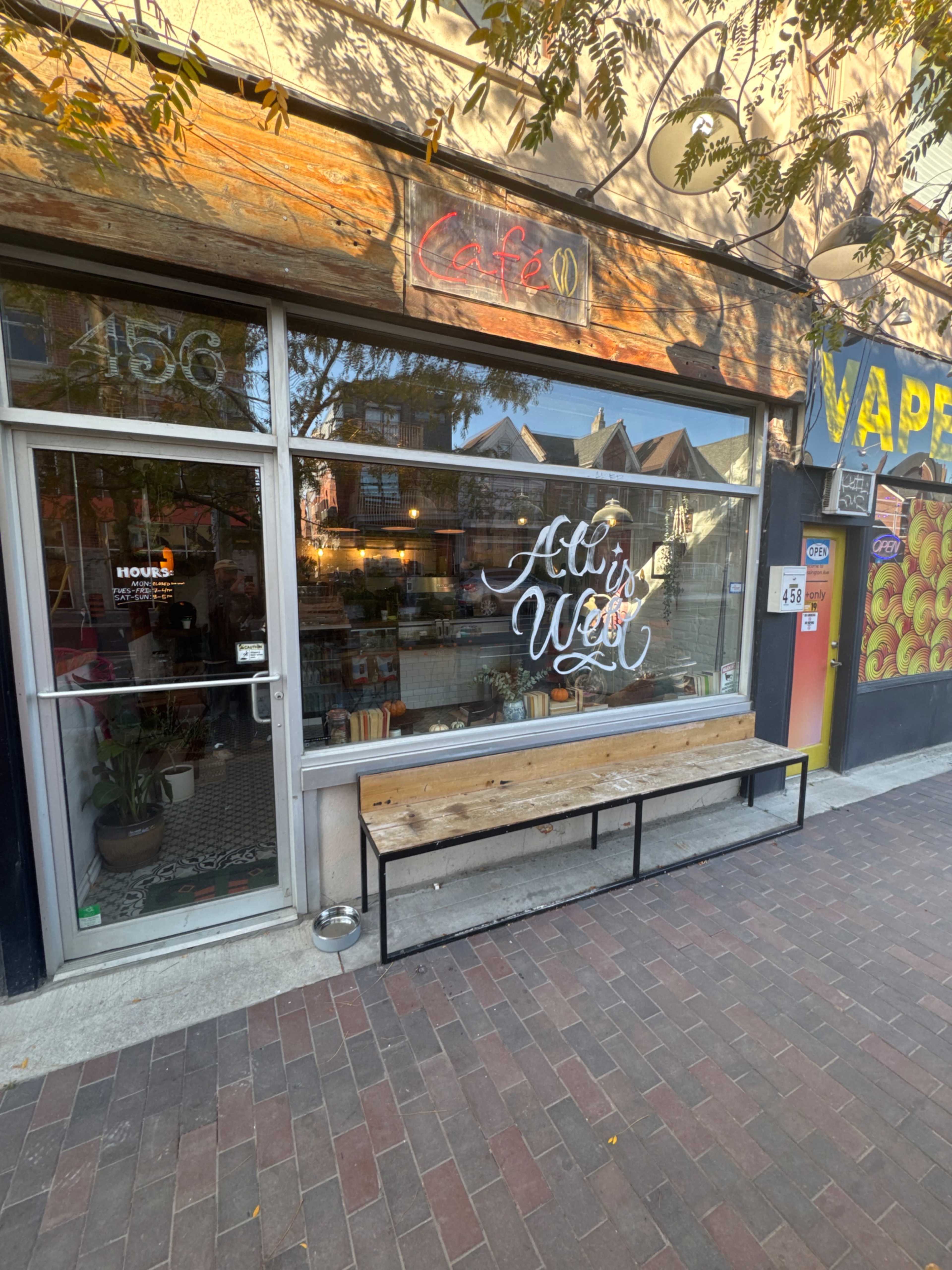 The image shows a café storefront with large glass windows displaying a handwritten sign that reads "All is Well," along with a wooden bench in front and various items visible inside.