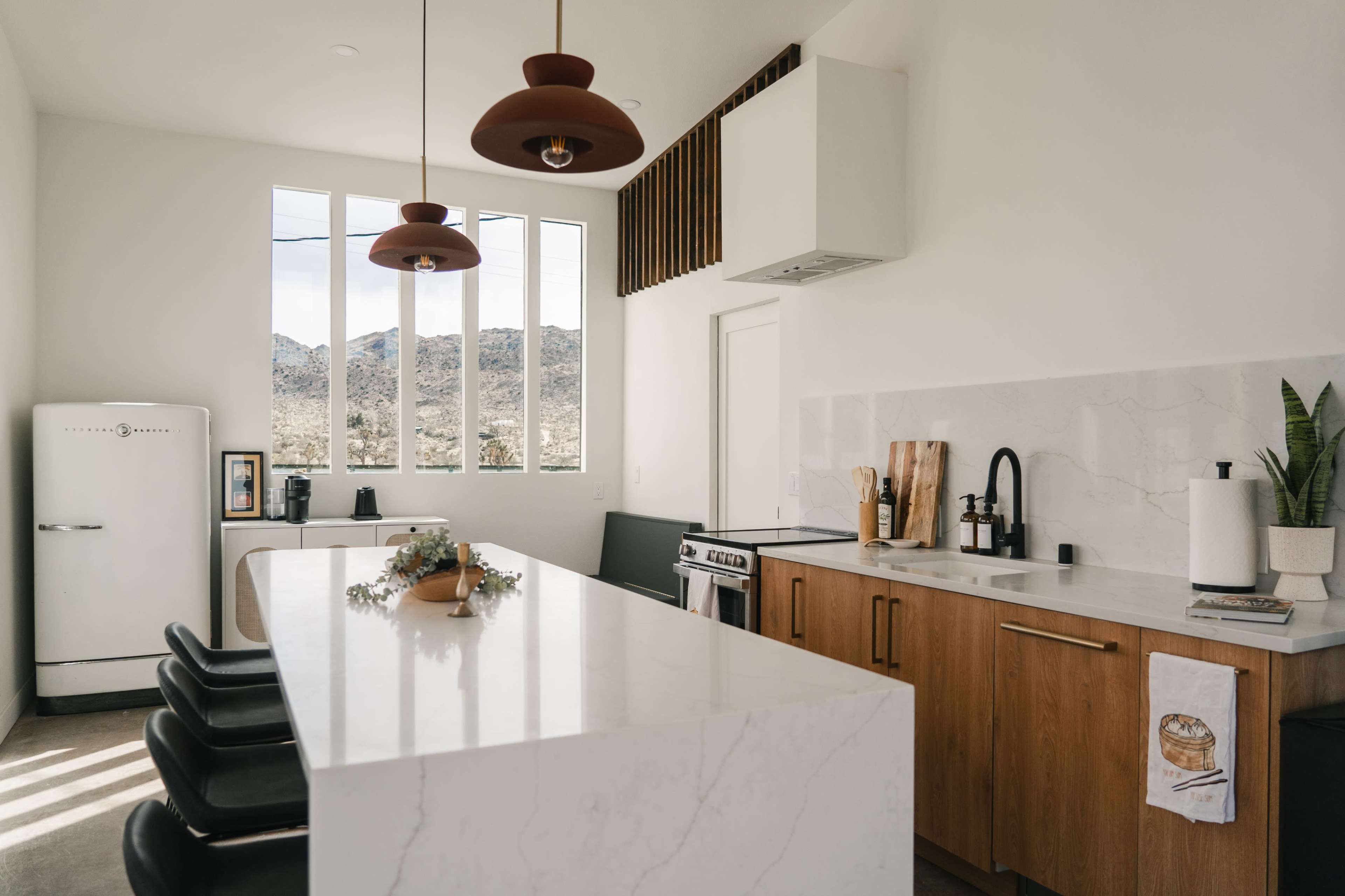 A modern kitchen features a central island with four black stools, wooden cabinetry, and three pendant lights hanging above.