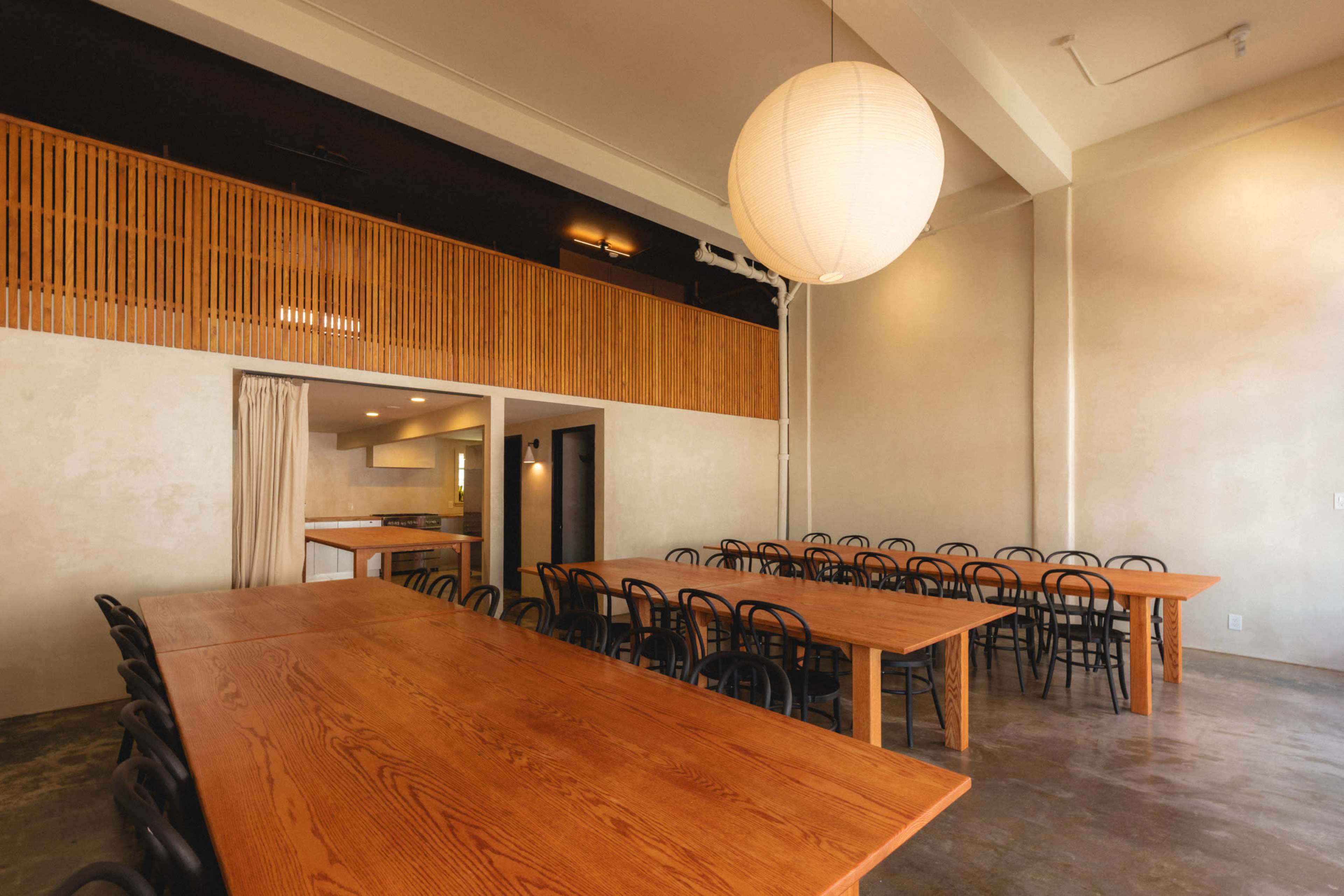 The image shows a spacious dining area with long wooden tables and black chairs, illuminated by a large pendant light.