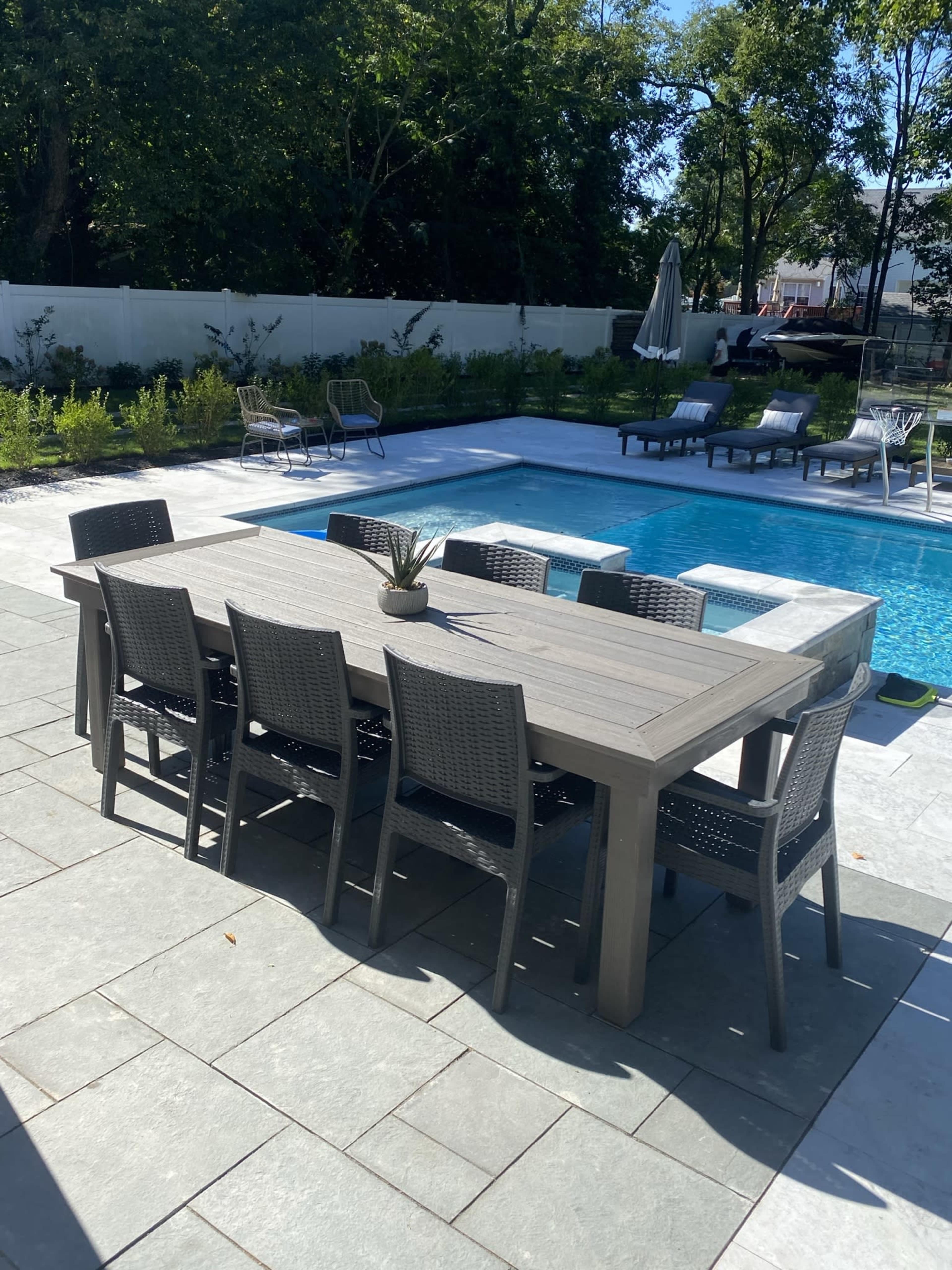 A large wooden dining table with eight chairs is positioned next to a swimming pool surrounded by greenery and lounge chairs.